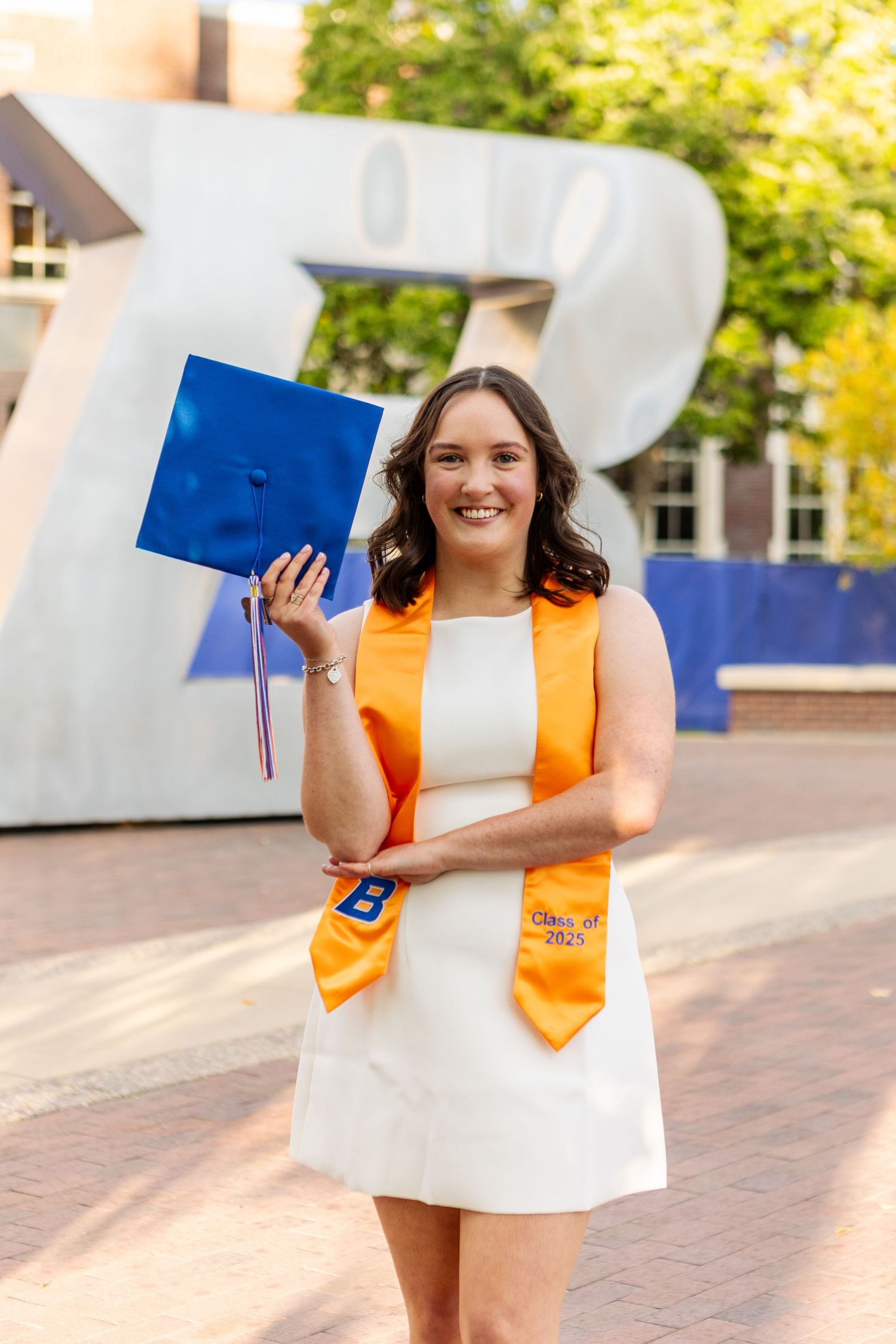 Graduate in white dress and orange stole celebrates with blue cap outdoors.