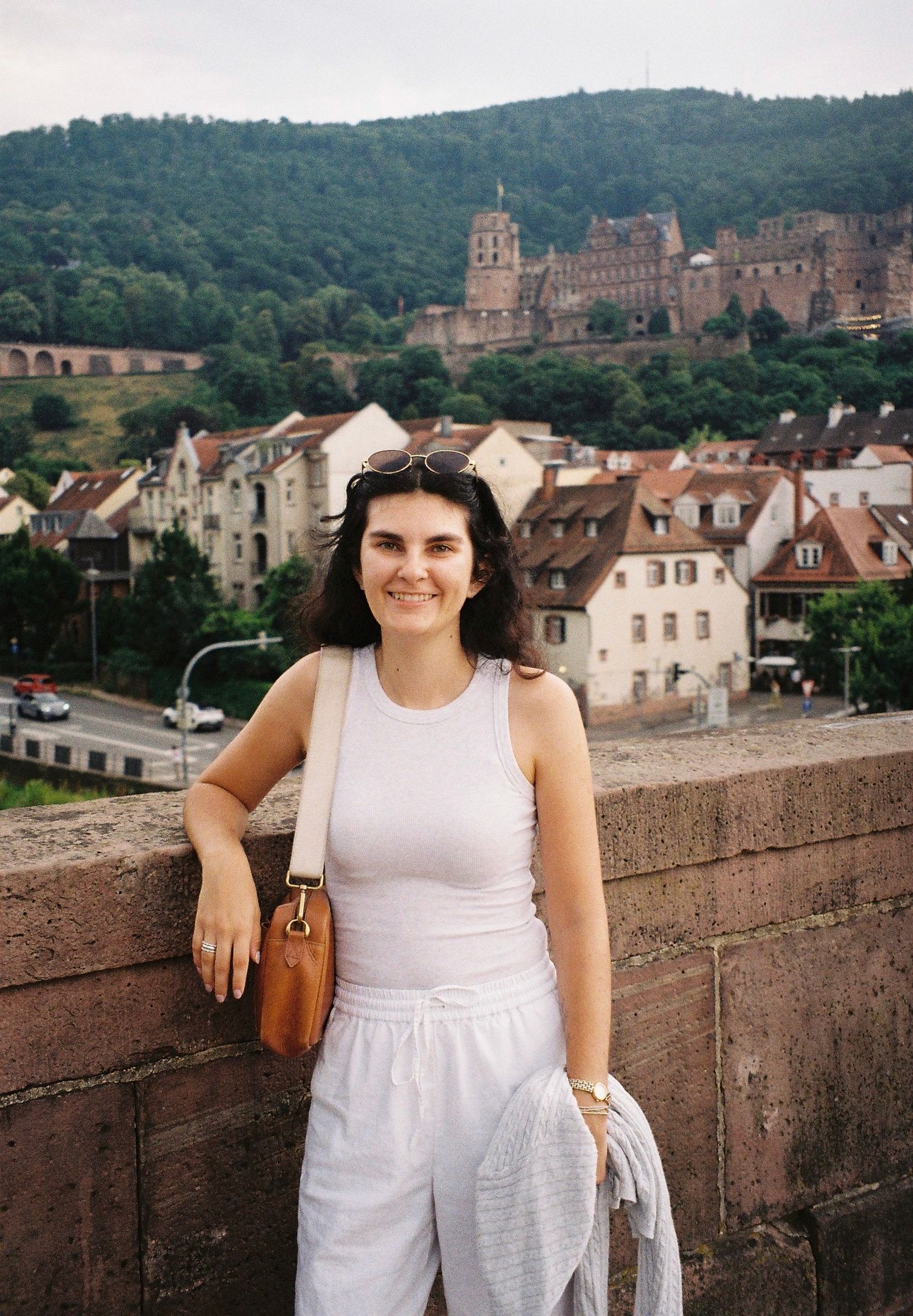 Tourist poses on stone wall with historic European castle and village in background.