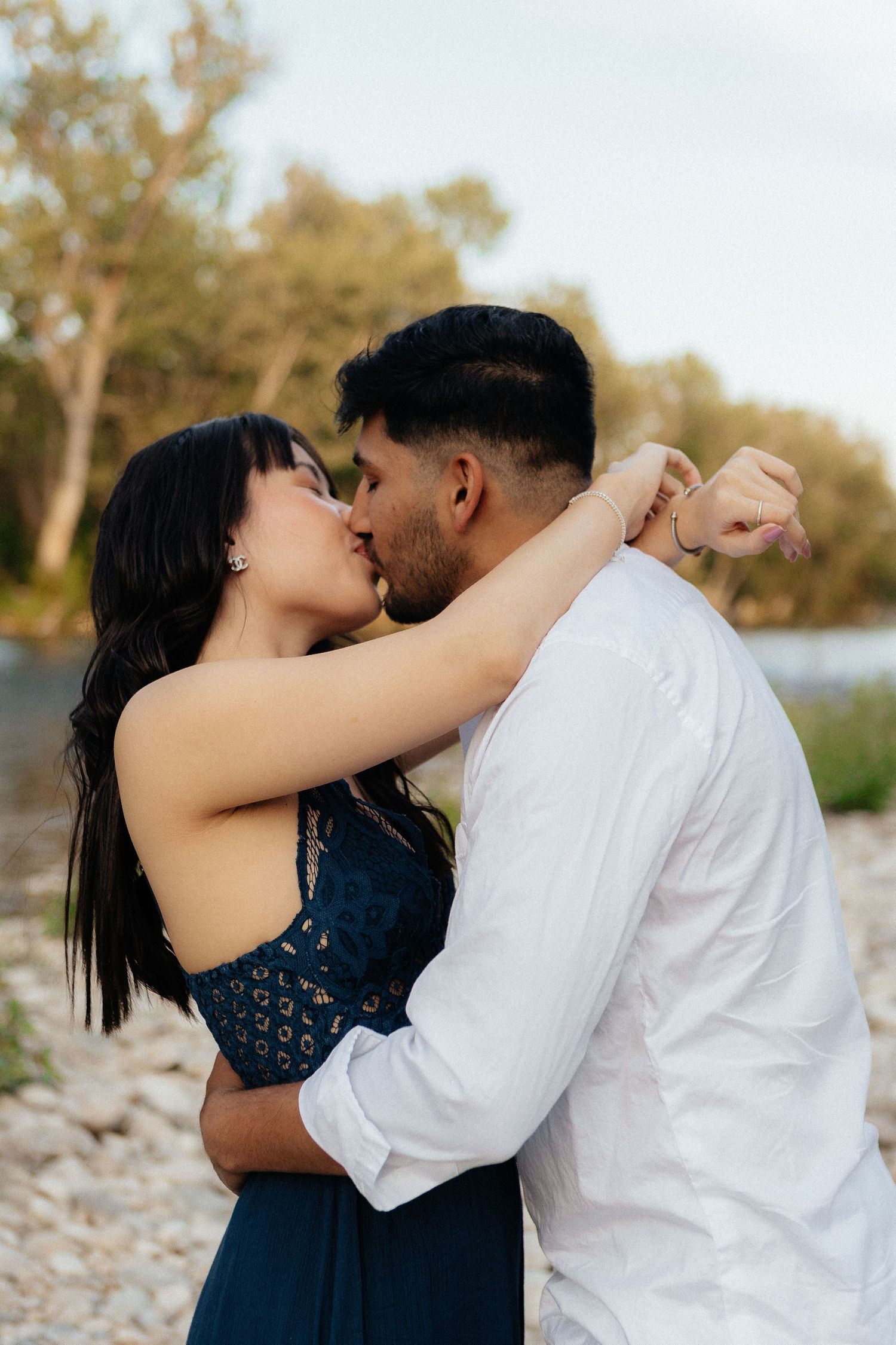 A romantic couple shares a tender kiss by a riverbank at sunset while embracing each other.