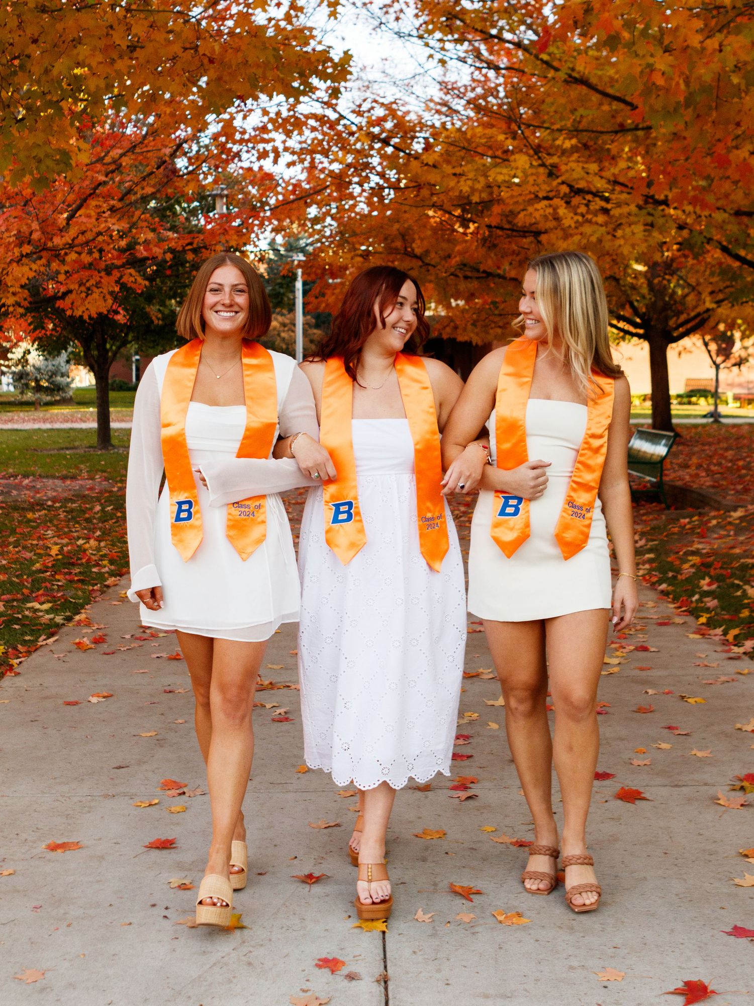 Graduates in white dresses and orange stoles walking under autumn trees with fallen leaves.