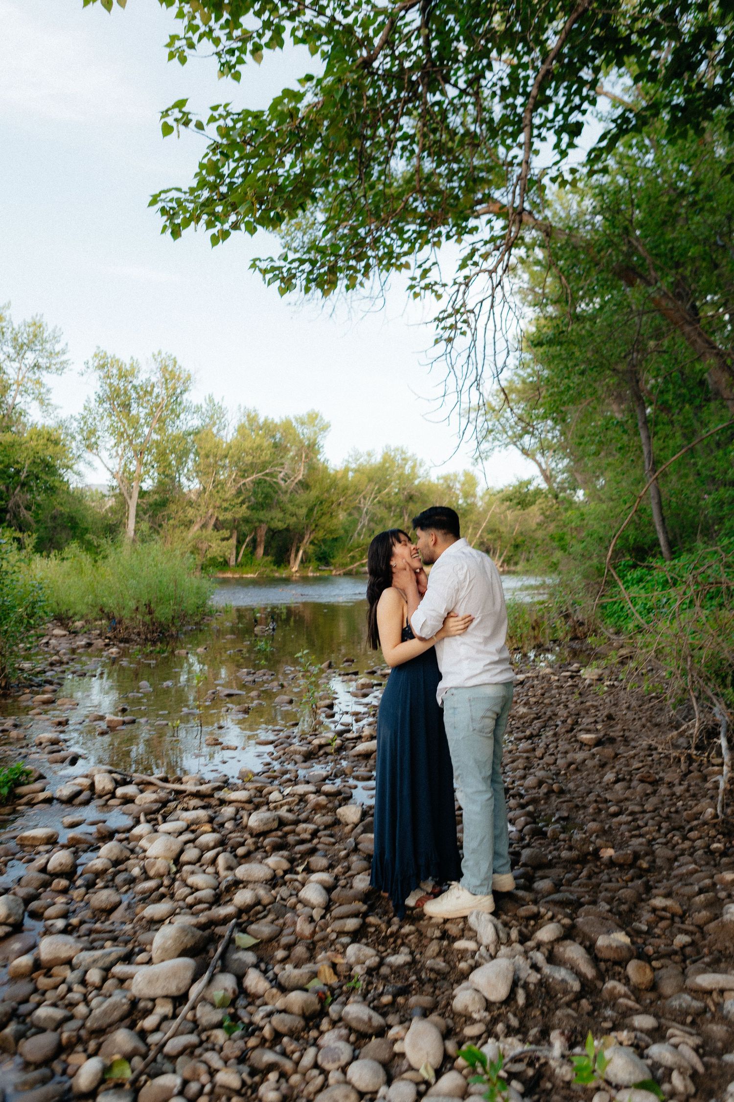 A couple shares a romantic moment on a rocky riverbank surrounded by green trees and flowing water.