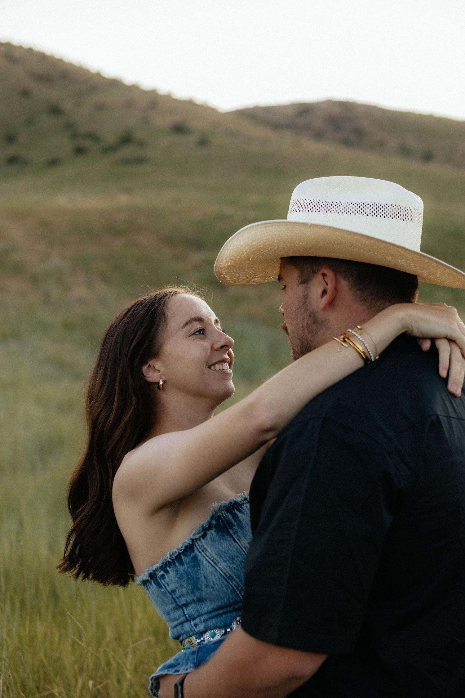 Romantic couple embracing in tall grass field at sunset with cowboy hat and casual western attire.