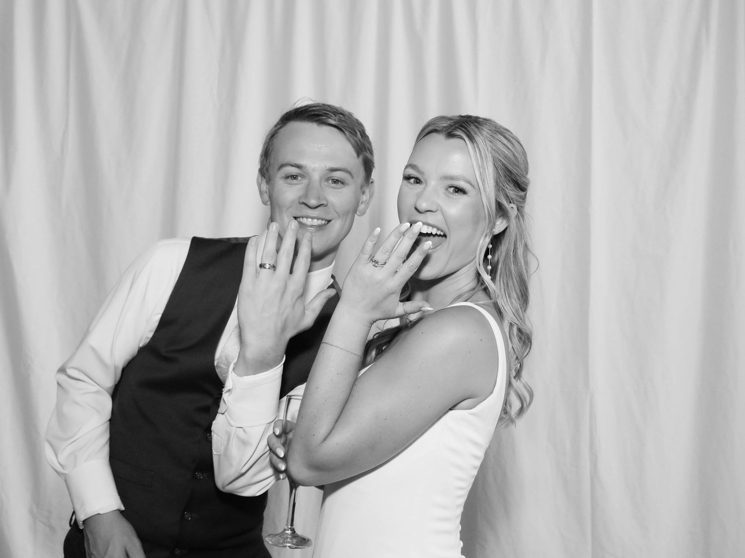 Black and white photo of newlyweds showing off wedding rings, smiling in a photo booth.