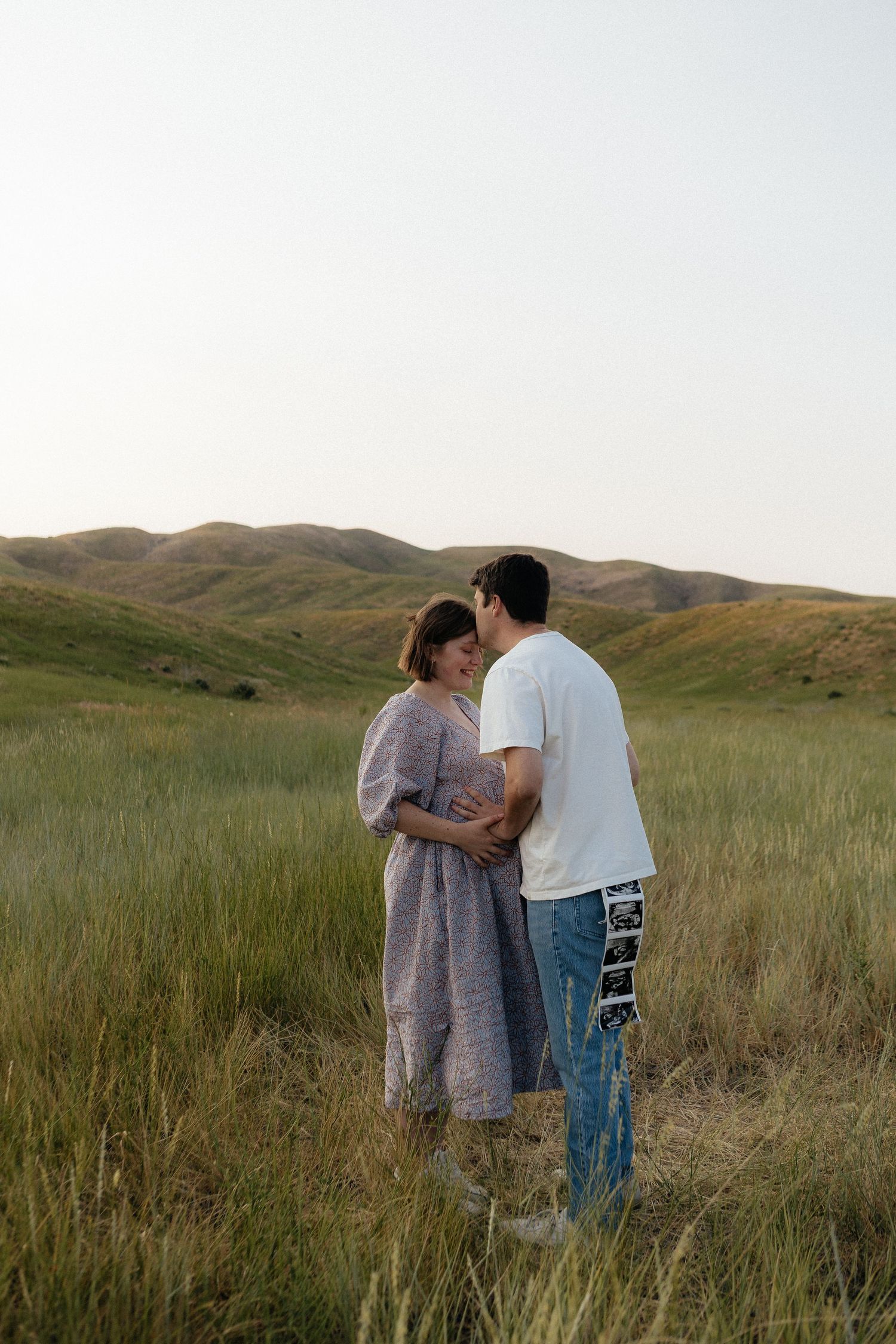 Couple stands close together in grassy field with golden hills in background during sunset.