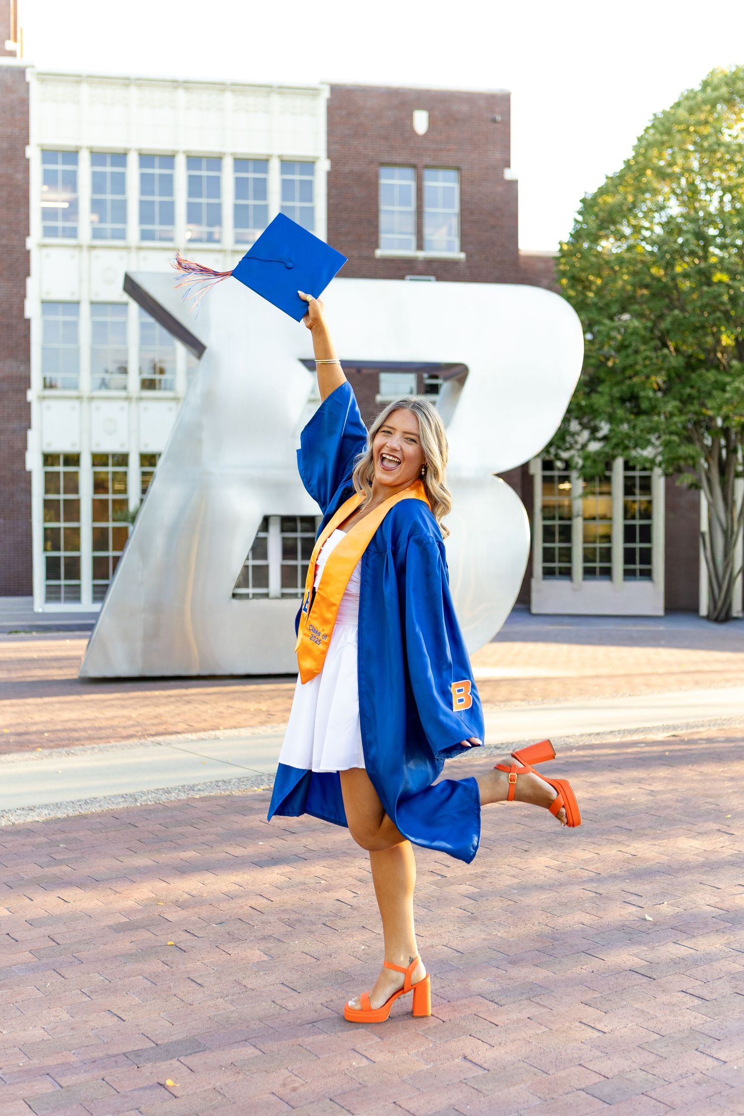 Graduate in blue cap and gown strikes playful poses on brick courtyard during photo session.