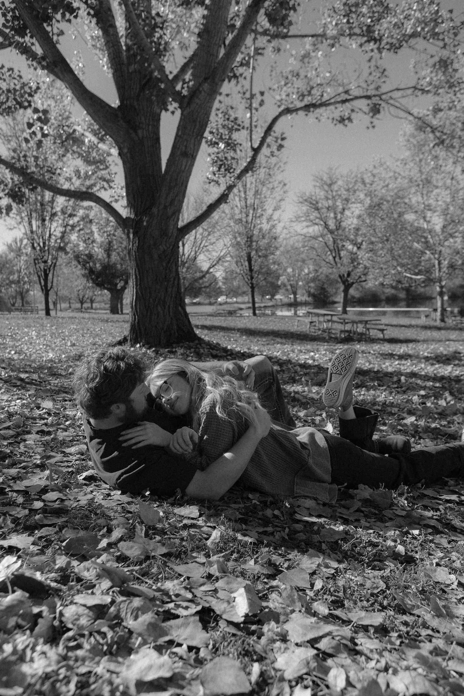 A couple lies together on fallen autumn leaves beneath a large tree in a park.