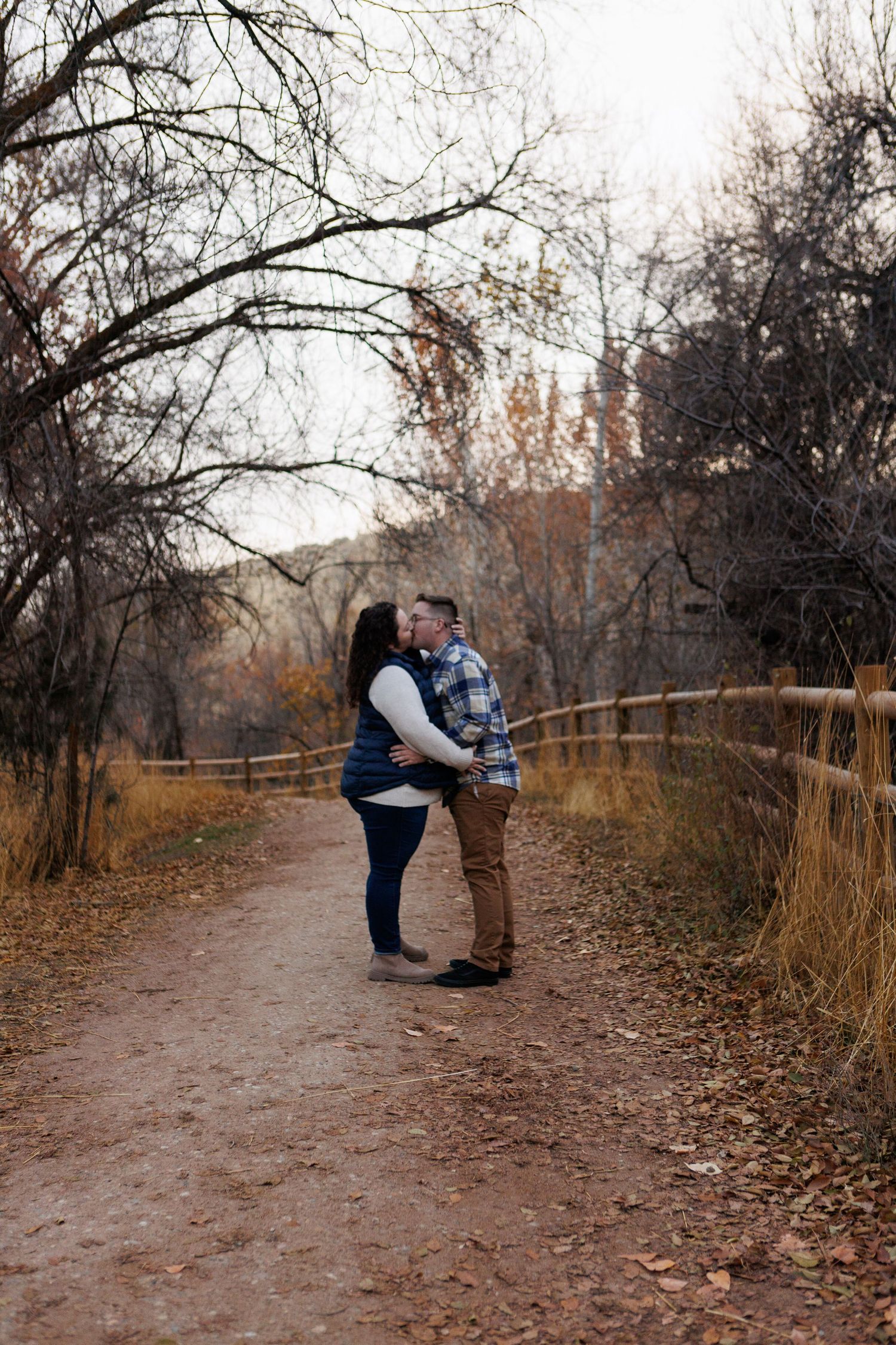 A couple shares a romantic moment on a dirt path surrounded by autumn trees and brush with a wooden fence beside them.