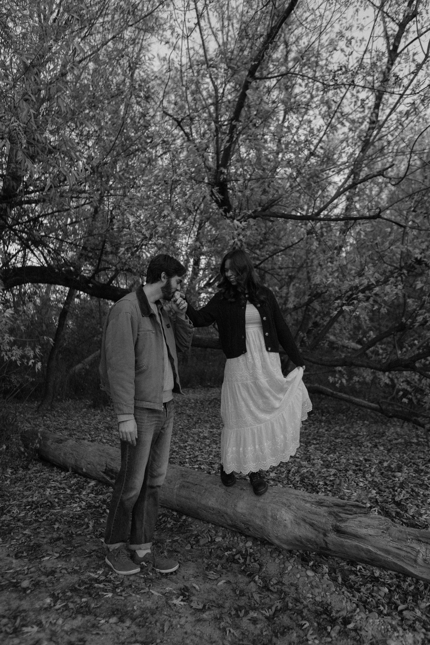 Couple explores wooded area with fallen leaves in black and white photography.