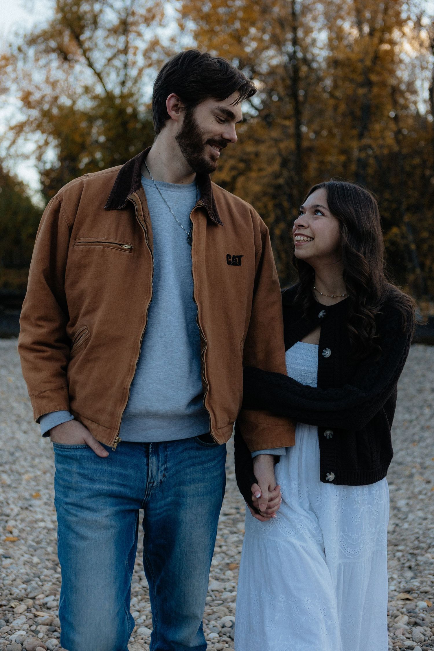 A couple in casual fall attire walks together on a leaf-covered path during autumn.