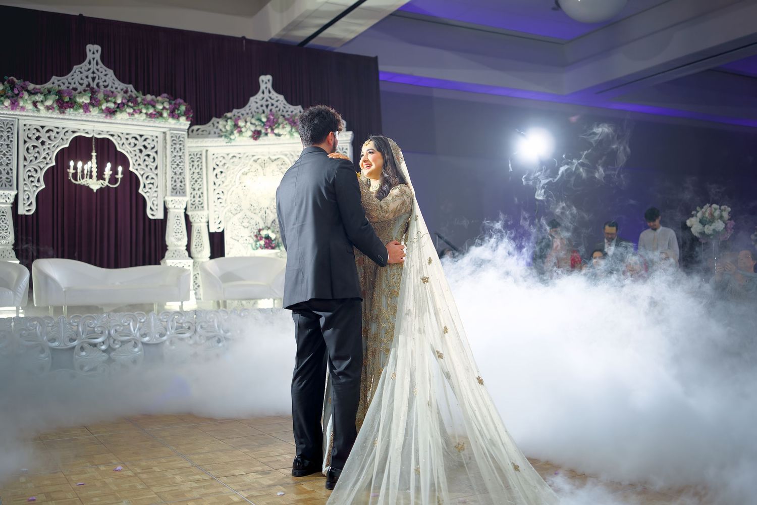 Newlyweds share their first dance on a misty dance floor illuminated by purple lights at an elegant wedding reception.