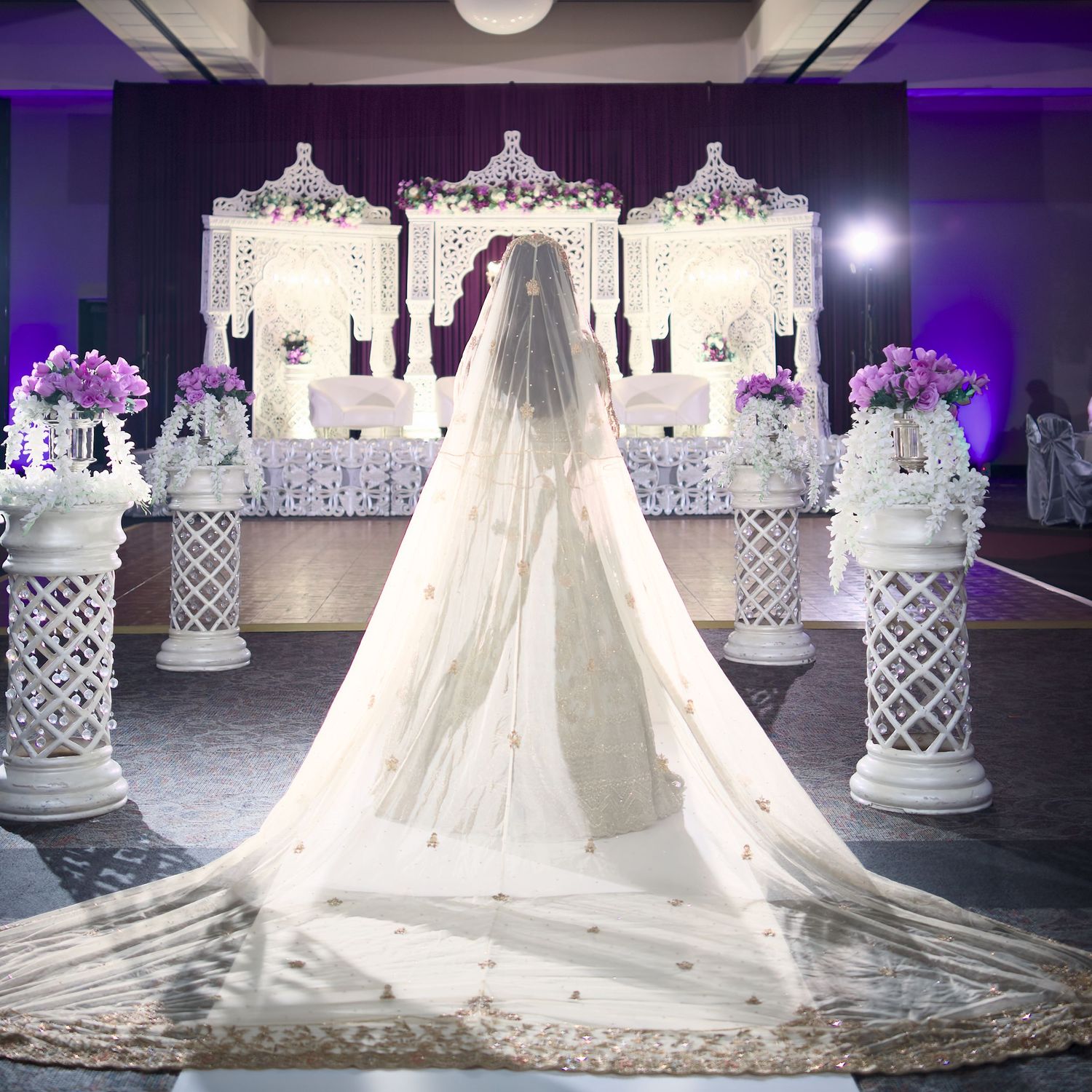 A stunning white wedding dress with cathedral veil displayed in a purple-lit venue with ornate white lattice decor elements.