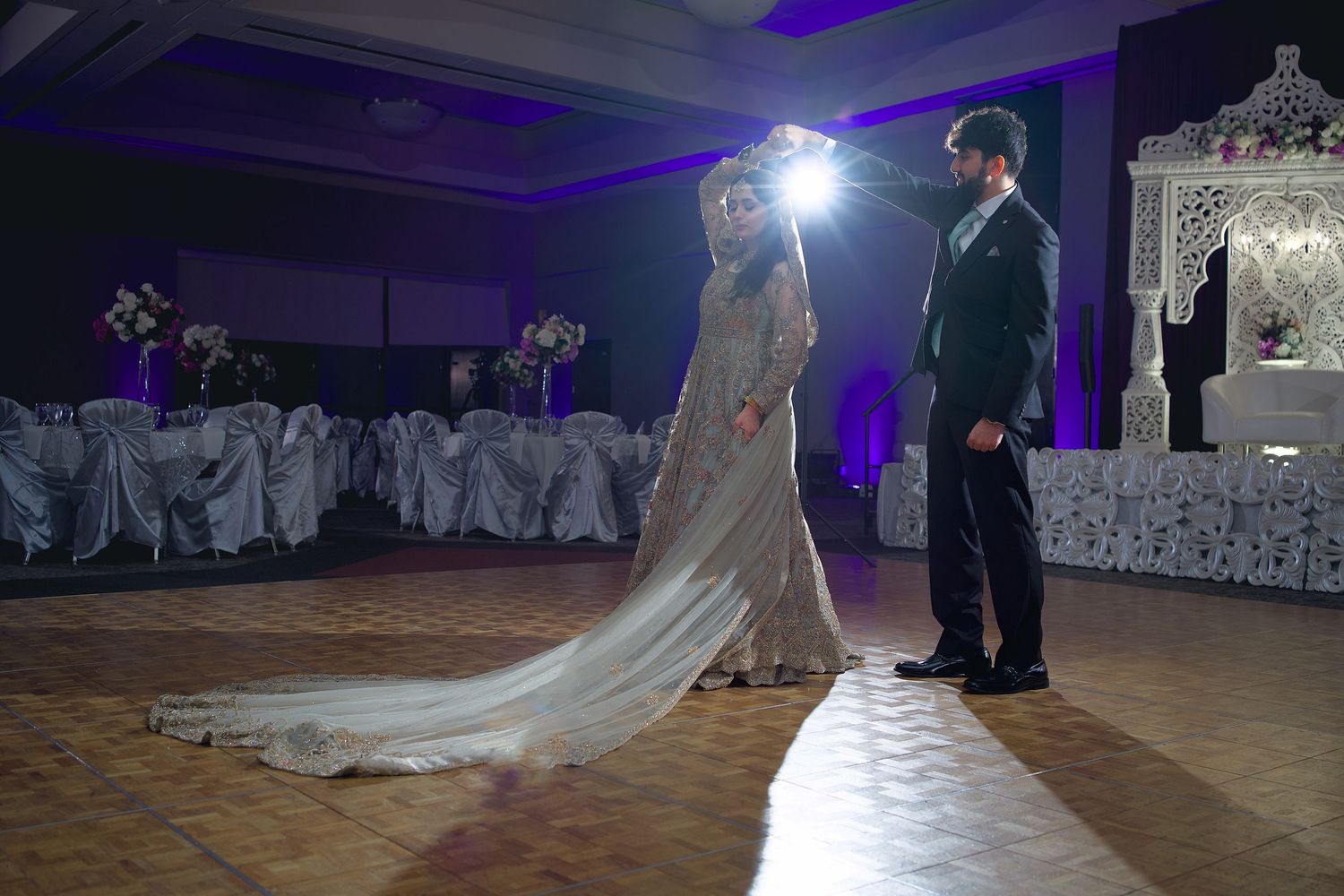 A couple performs their first dance at a wedding reception with dramatic purple lighting and a flowing white dress.