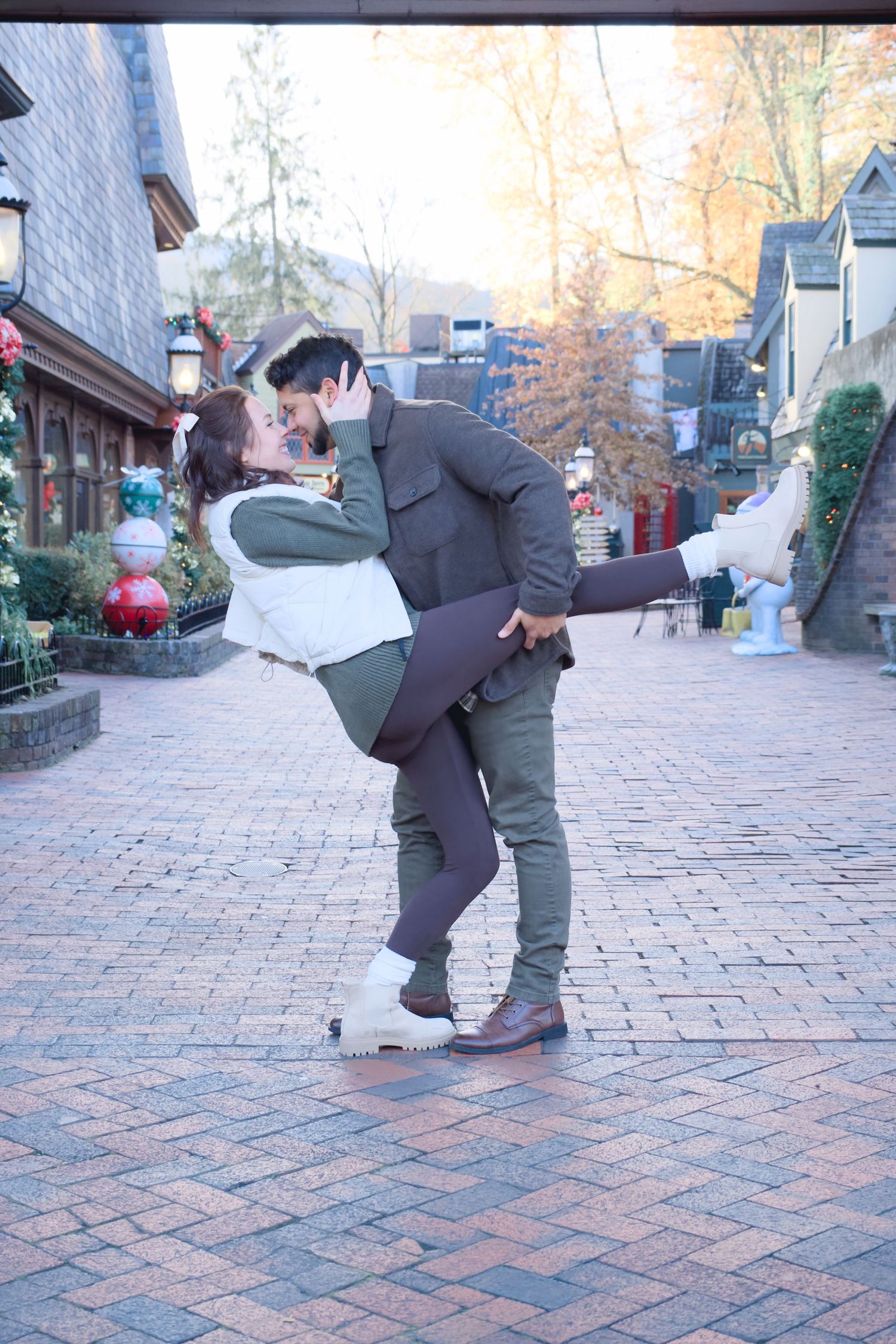 Playful couple dancing on brick pavement with holiday decorations in the background.