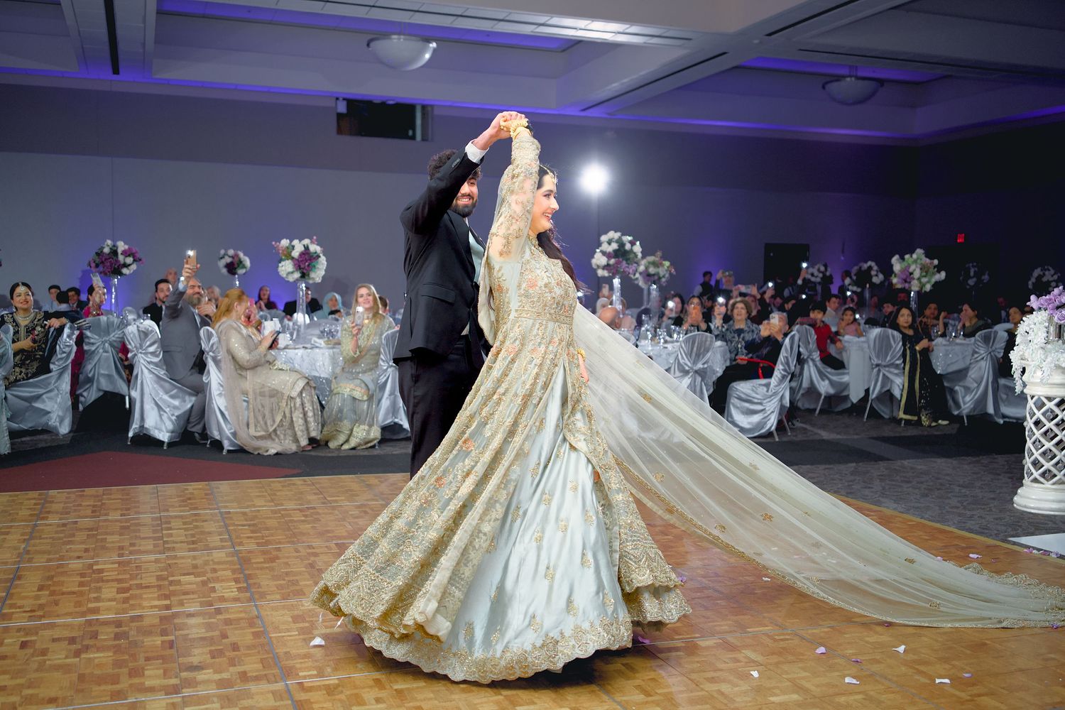 A couple performs their first dance at a wedding reception in a ballroom with guests watching from decorated tables.
