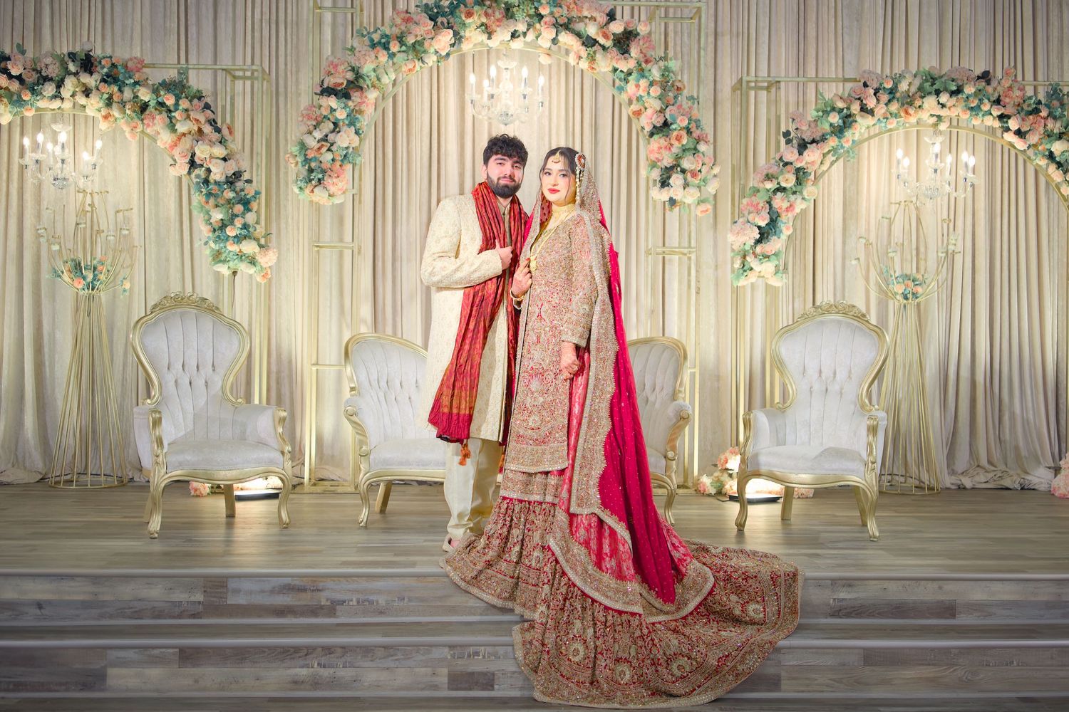 Traditional South Asian wedding couple poses under floral arch with elegant white throne chairs and draped backdrop decor.