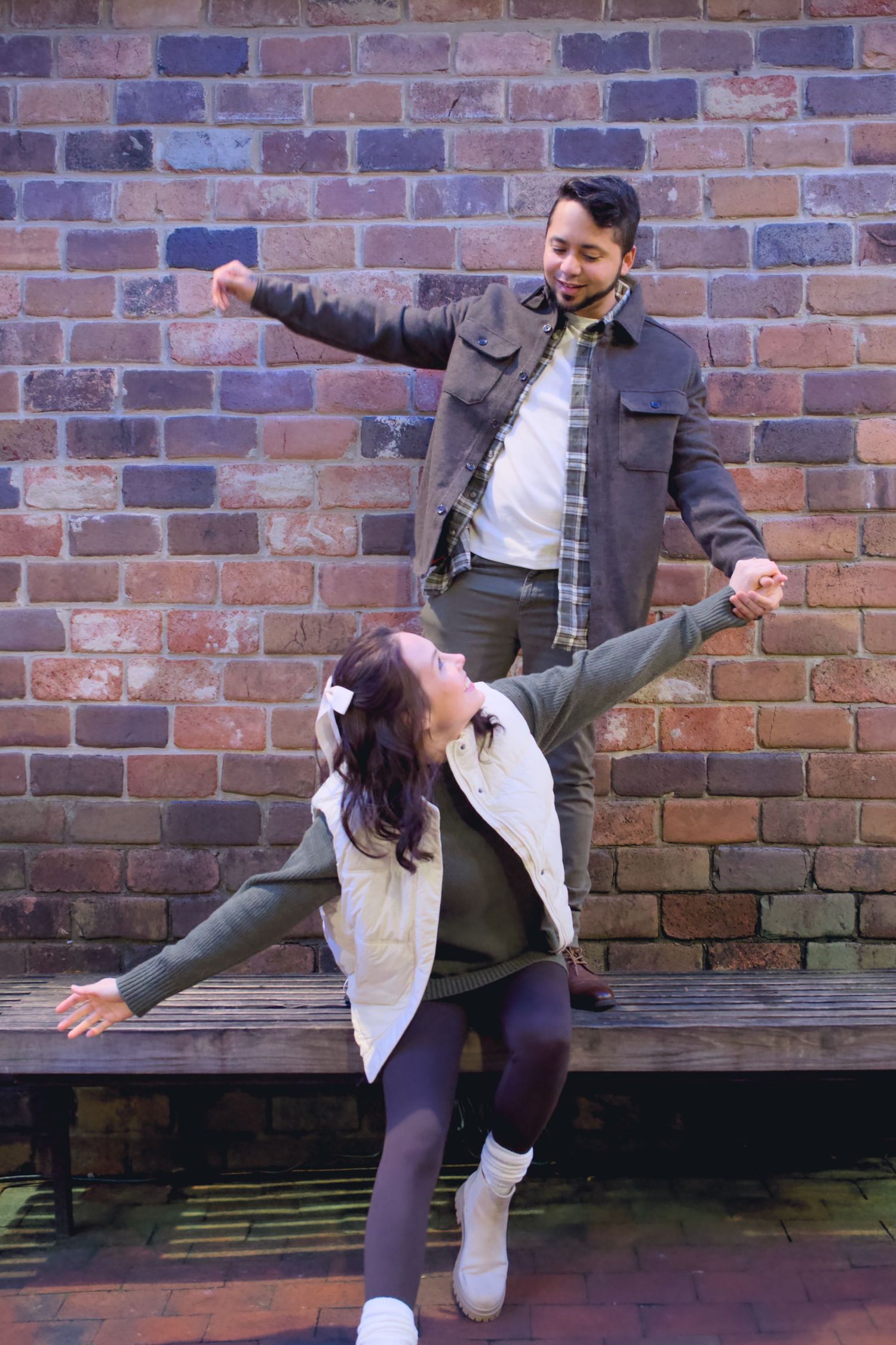 Couple performs playful dance moves against a brick wall backdrop.