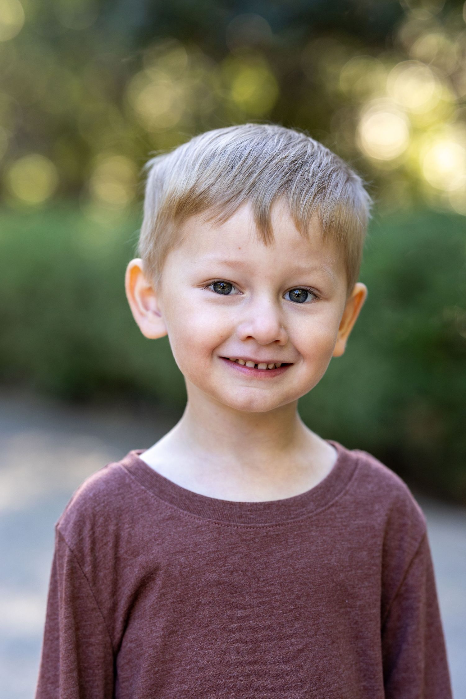 A young child in a brown sweater smiles at the camera in an outdoor setting.