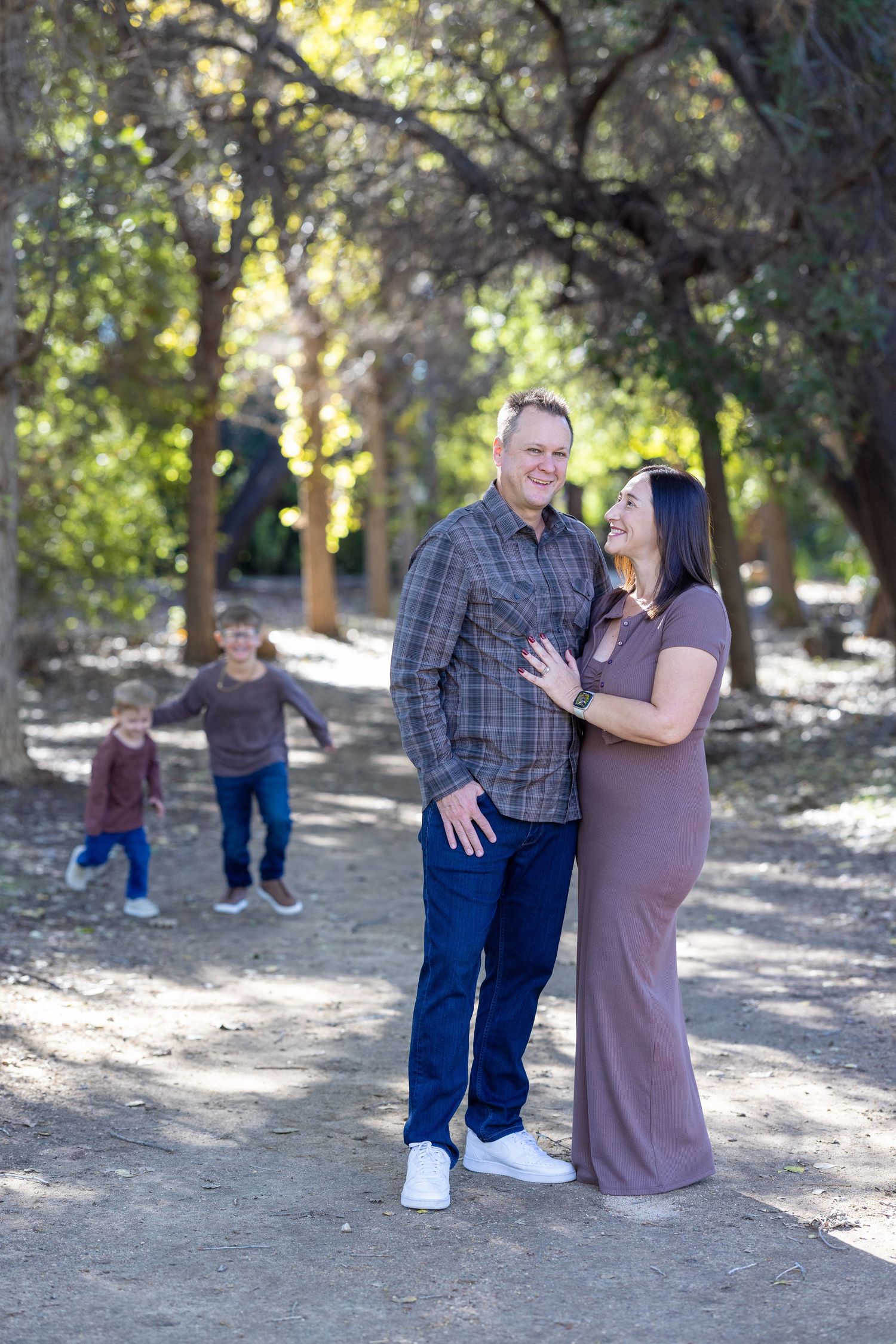 A couple shares an intimate moment on a wooded path while children play in the background.