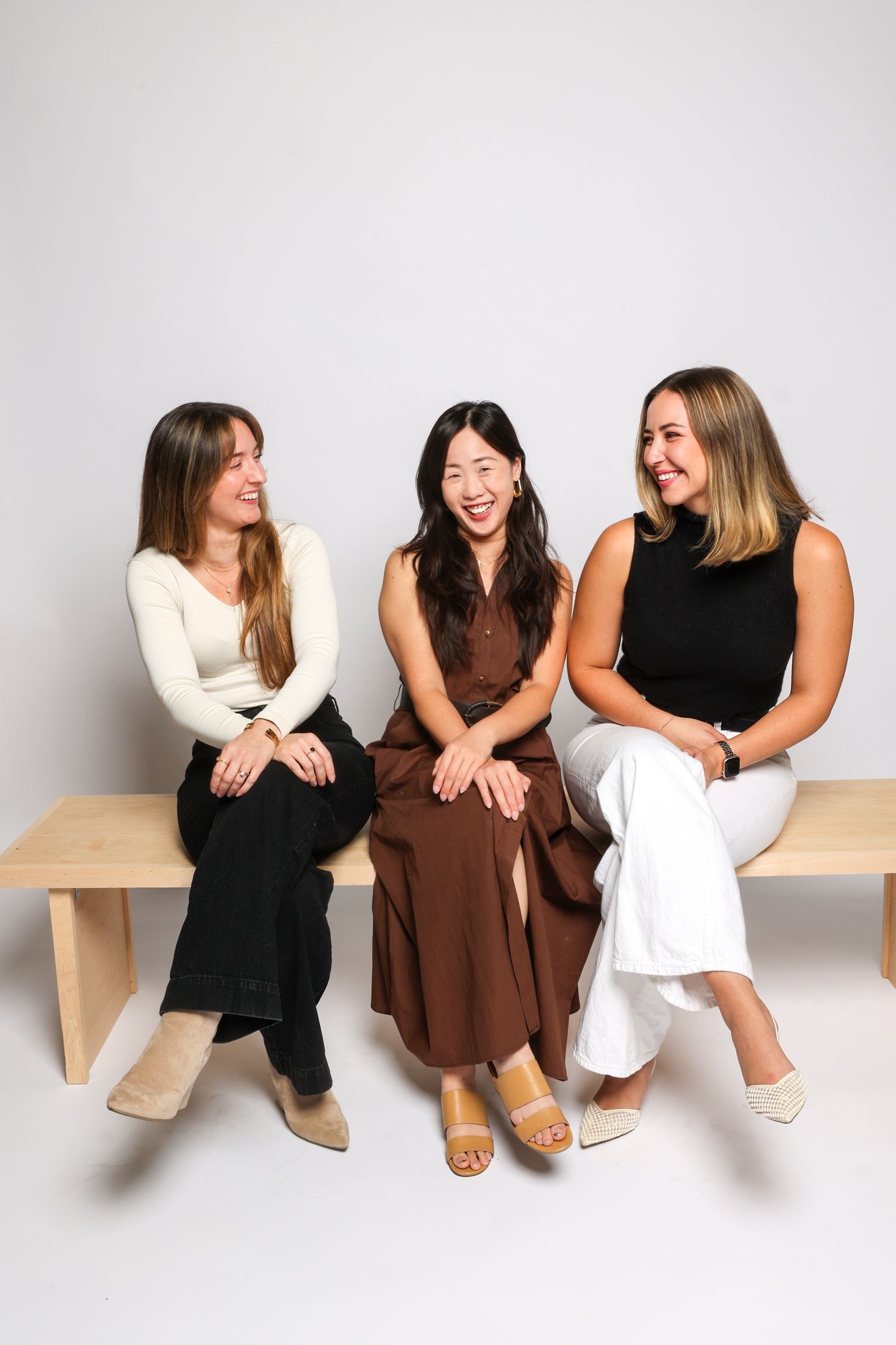 Three women sitting on bench in casual dresses sharing candid laughing moments.