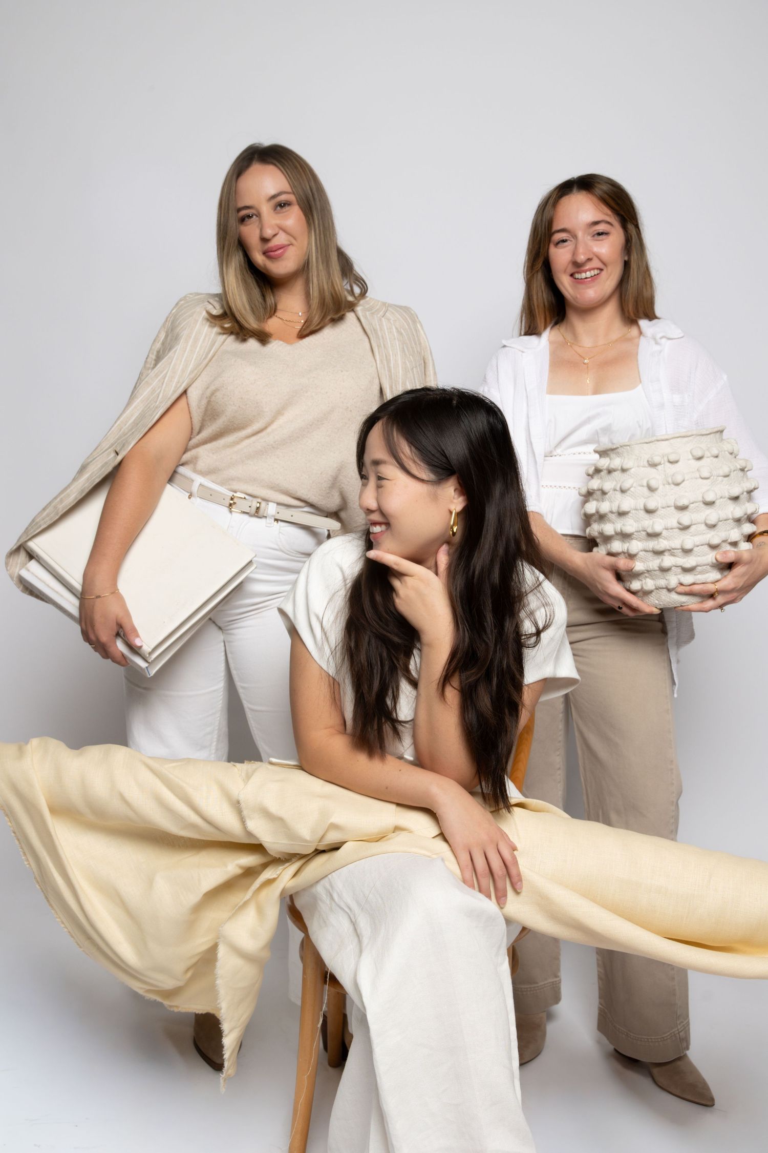 Women in neutral tones posing playfully with fabric swatches in studio setting.