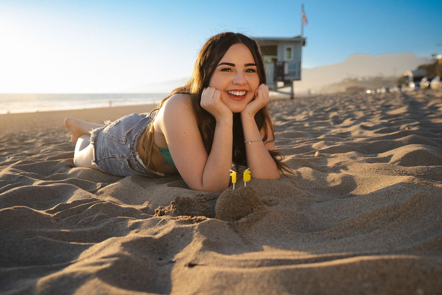 A person lies on beach sand while smiling at the camera during golden hour.