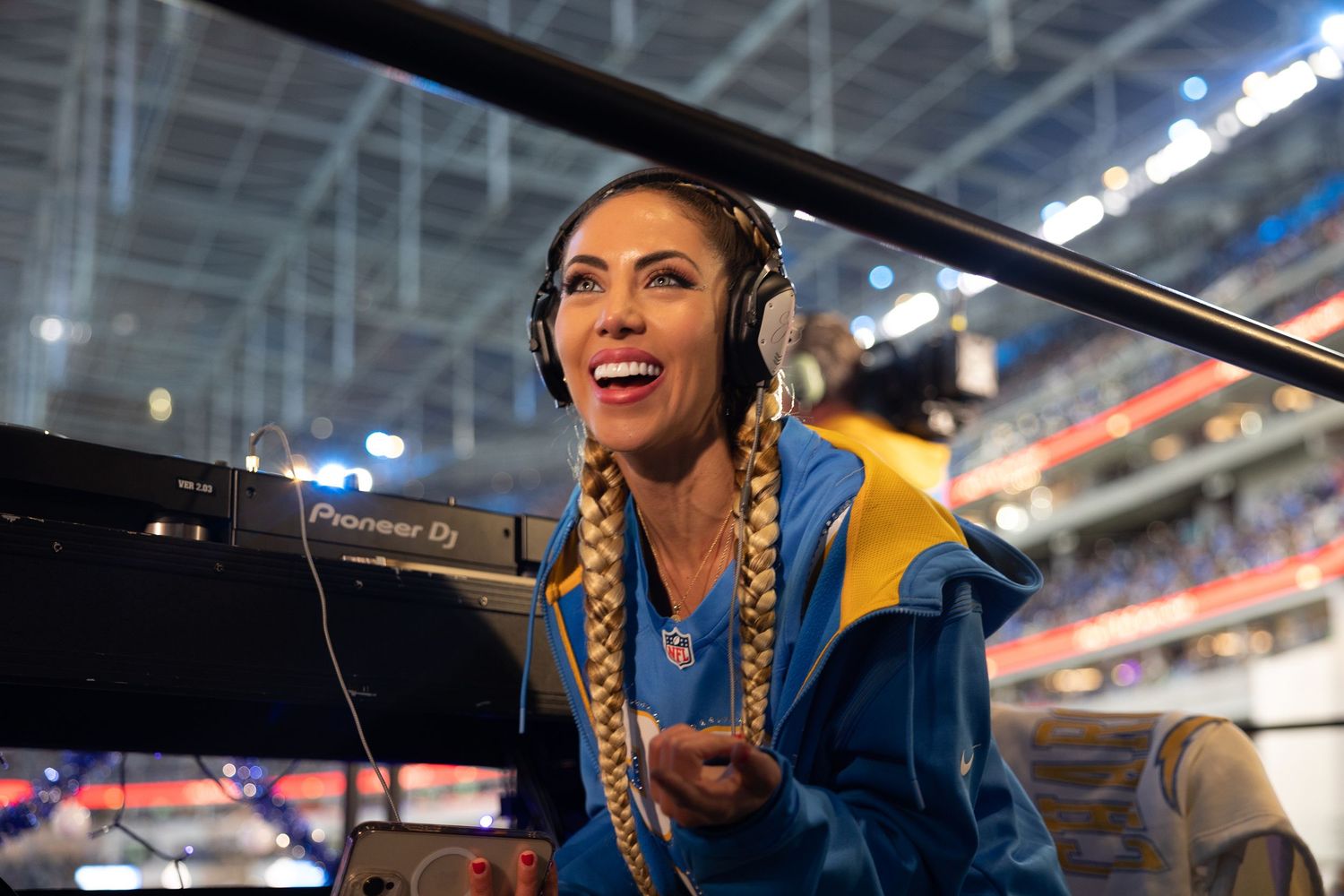 A sports announcer with braided hair and headphones broadcasts enthusiastically from a stadium booth.