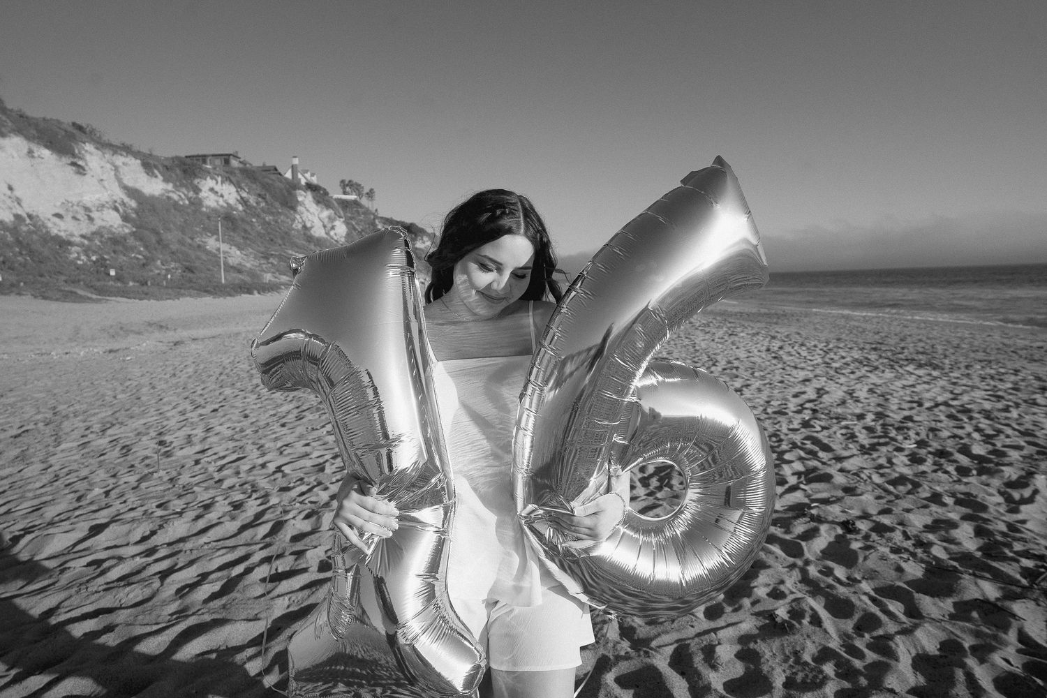 Someone poses with large silver number balloons on a sandy beach.