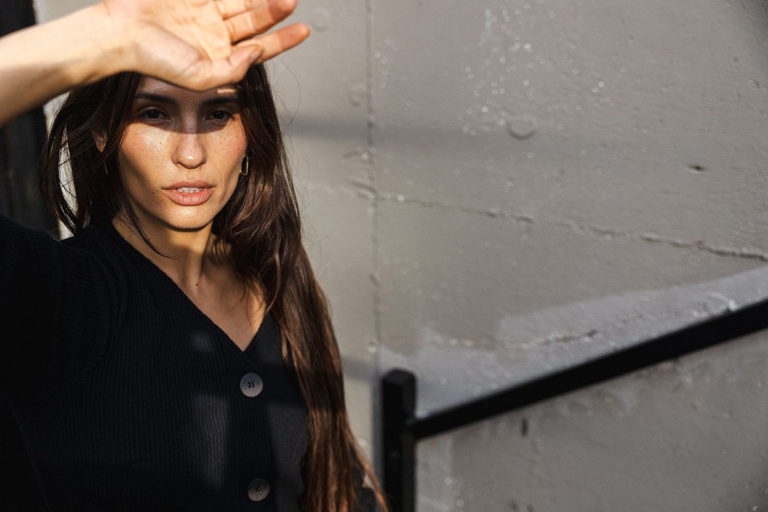 Model in dark outfit poses dramatically against grey wall.