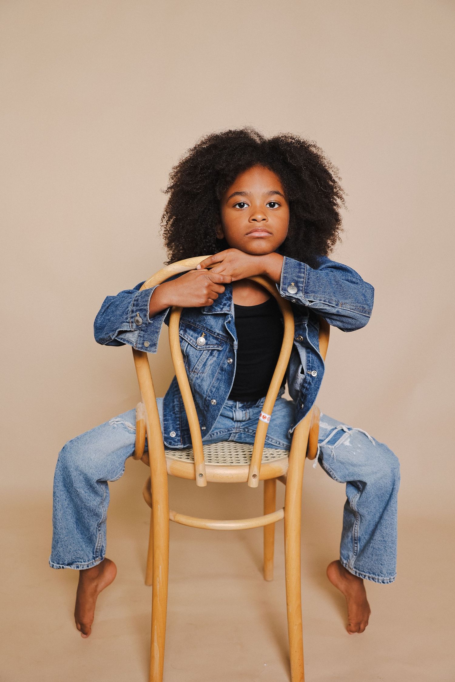 Child in denim outfit poses creatively on wooden chair against beige backdrop.