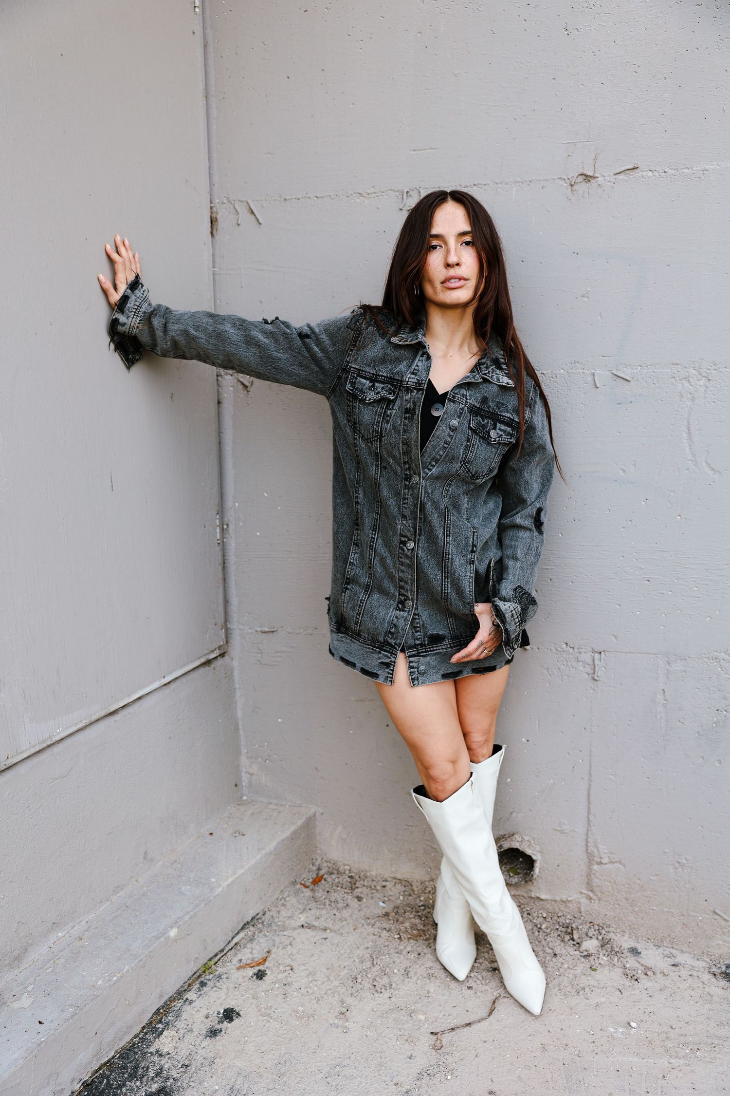 Fashion model wearing denim jacket and white boots poses against white wall.