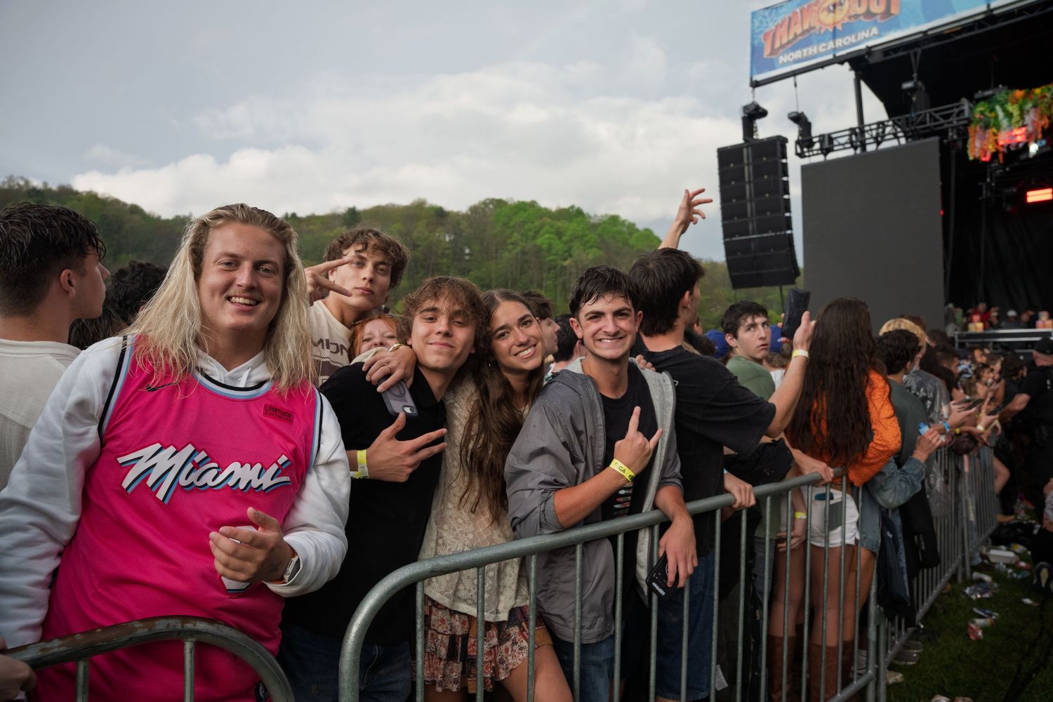 Smiling young festival fans pose together at metal barrier during outdoor music concert on a cloudy day.