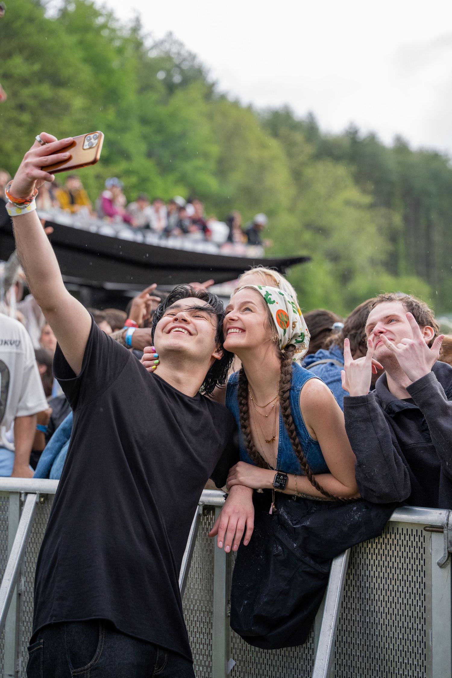 Man and woman smiling taking selfie together at outdoor music festival near metal barrier.