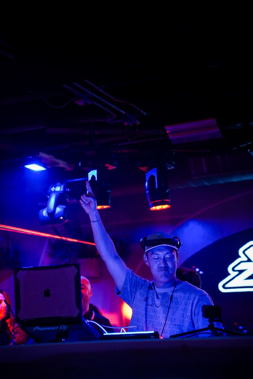 DJ raising one hand while performing at a nightclub with blue and red laser light effects.
