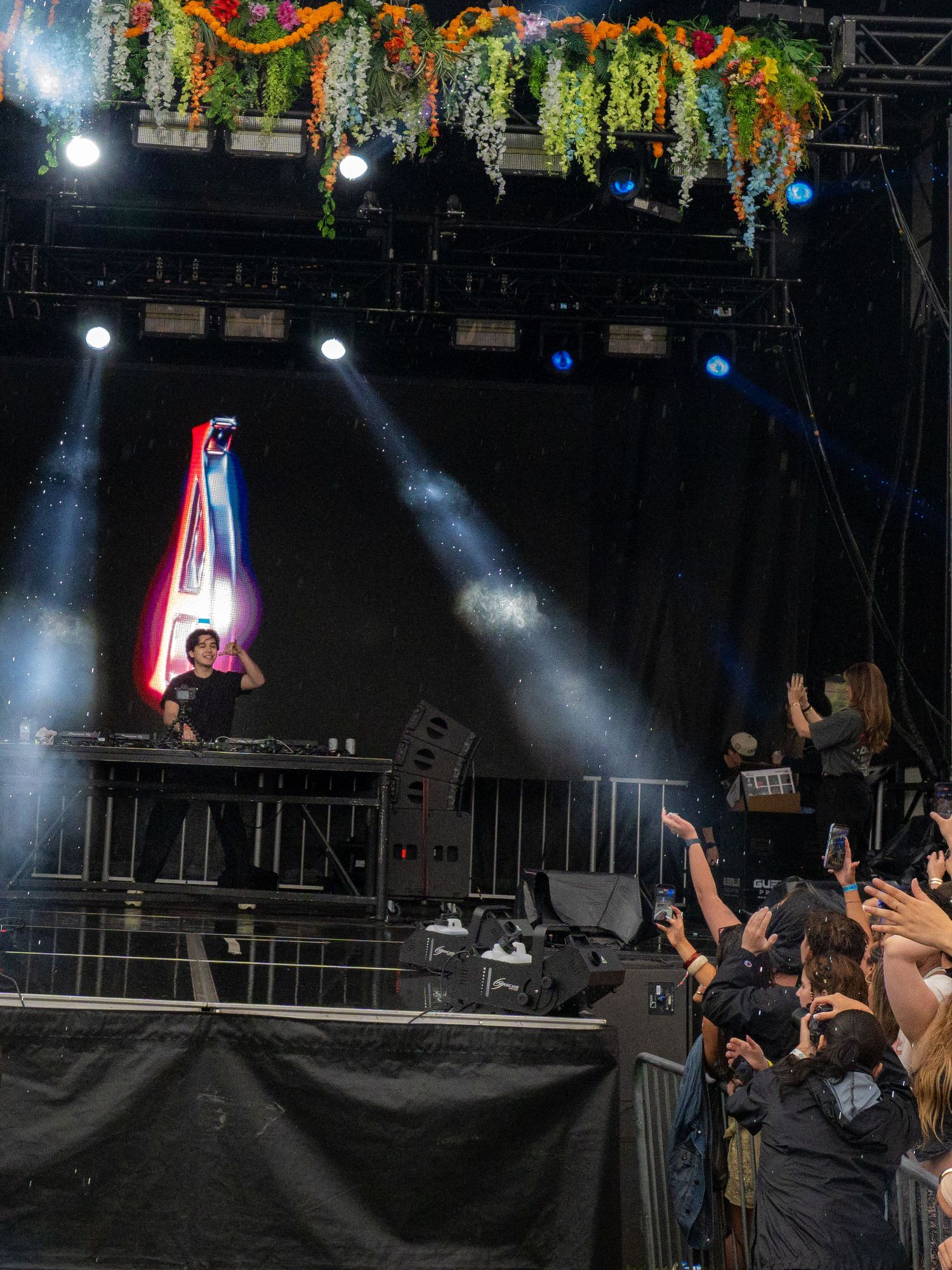 A DJ performs on a decorated stage with colorful lighting at an outdoor music festival.
