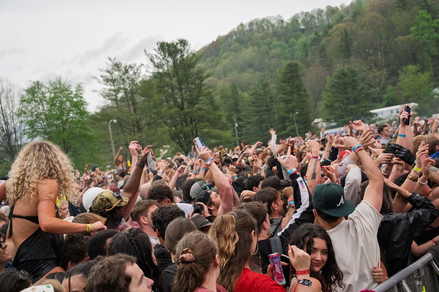 Large enthusiastic crowd with raised hands packed together at an outdoor music festival surrounded by green hills.