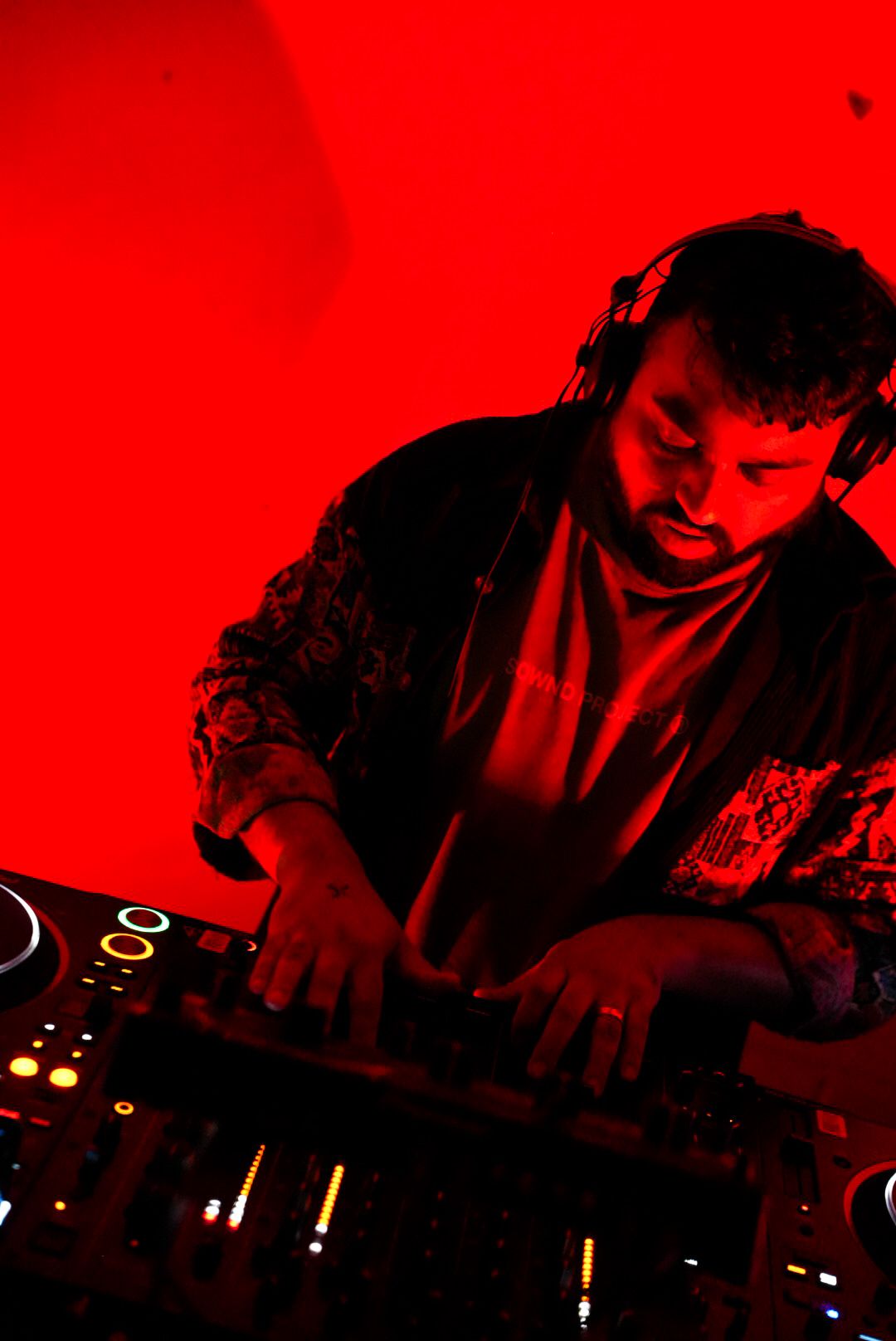 Male DJ wearing headphones focuses on mixing tracks under vivid red club lighting.