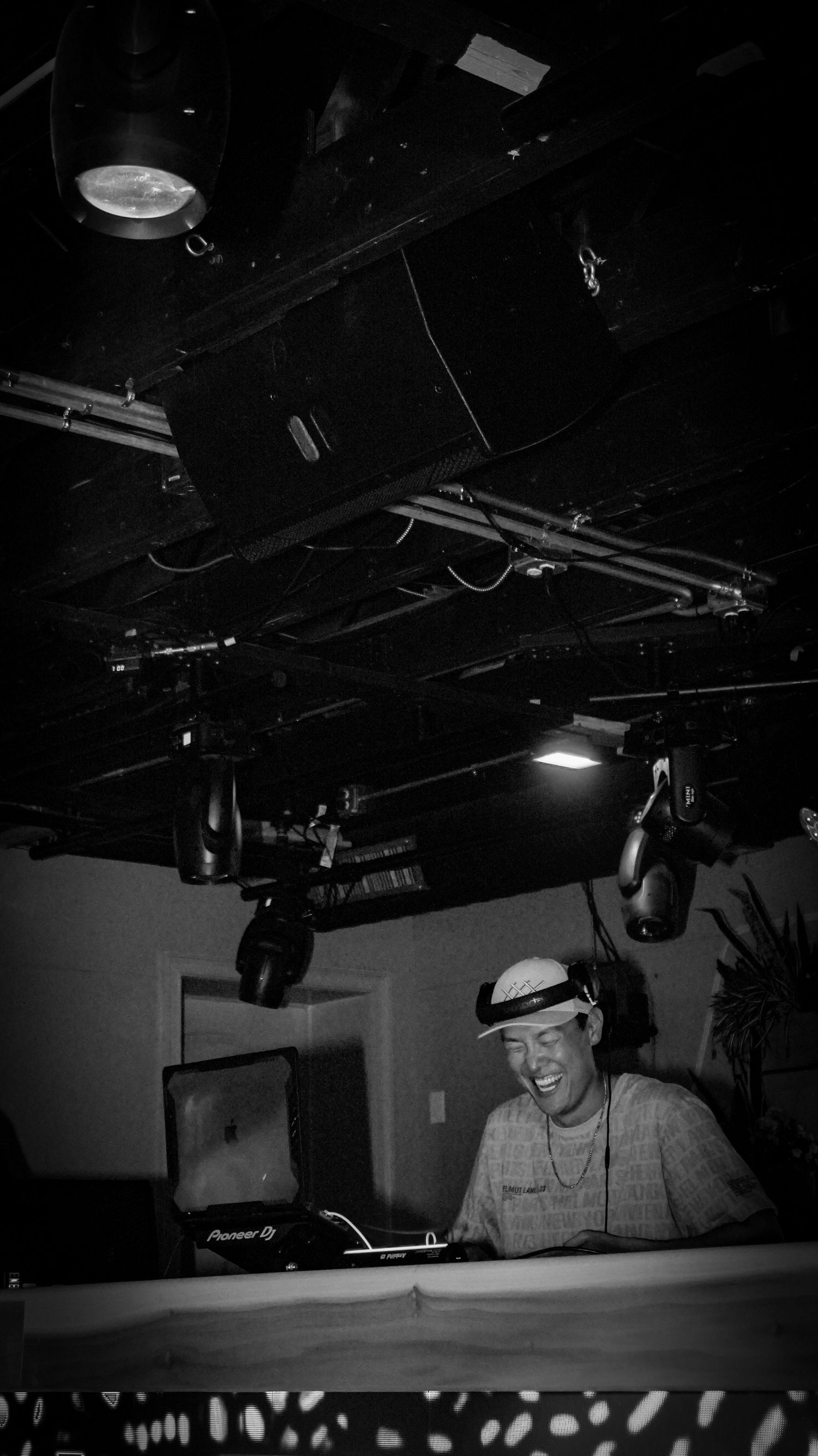 Black and white photo of a smiling DJ wearing a cap working at Pioneer DJ decks in a nightclub.
