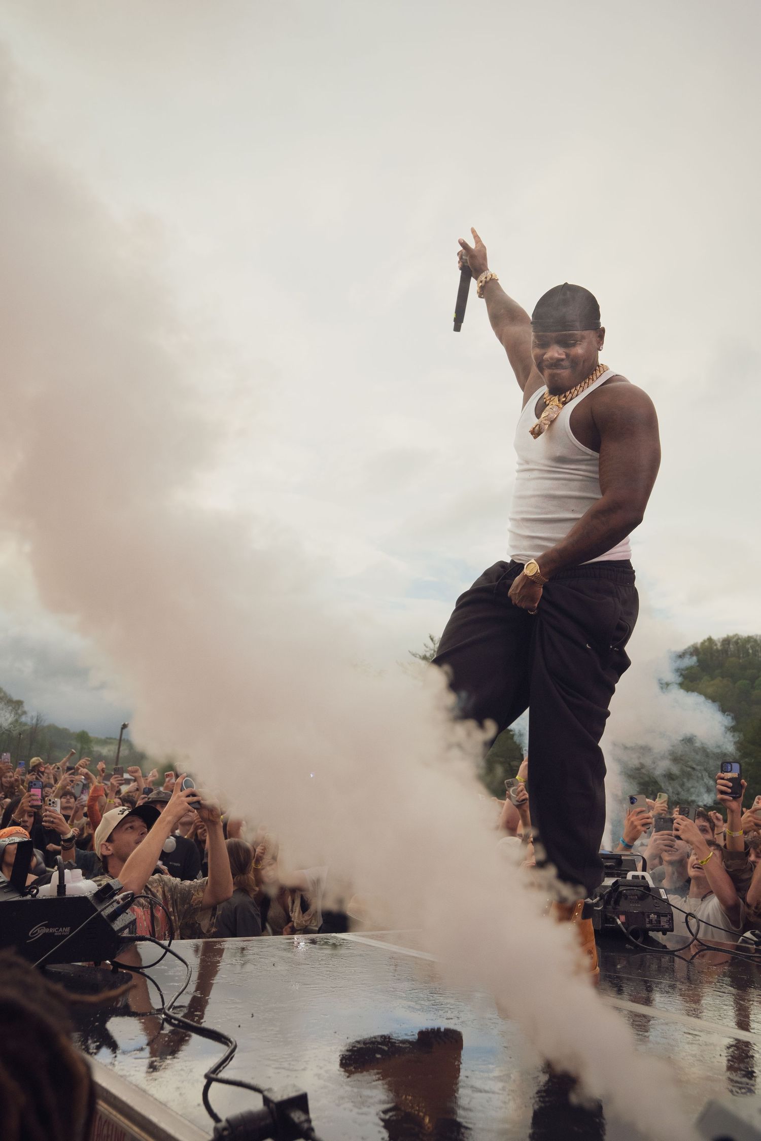 Rapper poses dramatically amid pink smoke on stage edge at outdoor music festival.