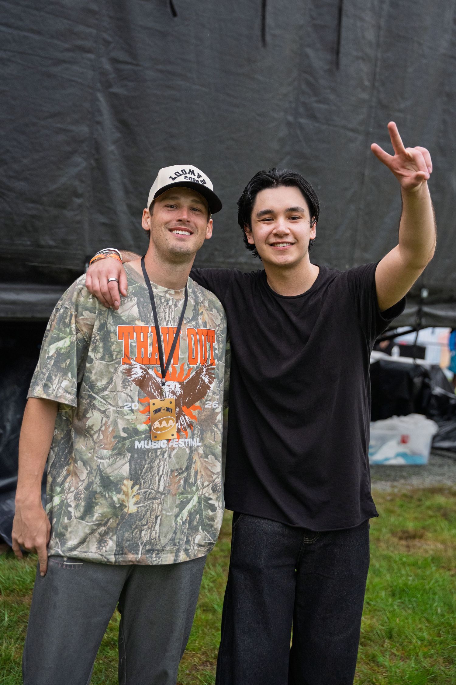 Two young men pose backstage at Thank You Music Festival, one flashing peace sign.