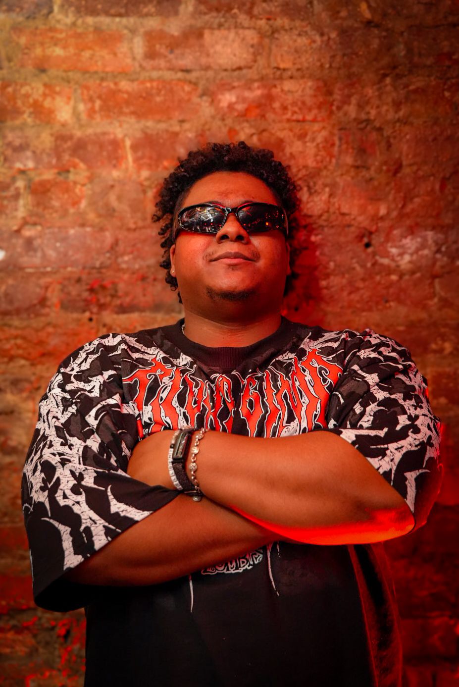 Confident young man in Thrasher shirt with arms crossed against brick wall.
