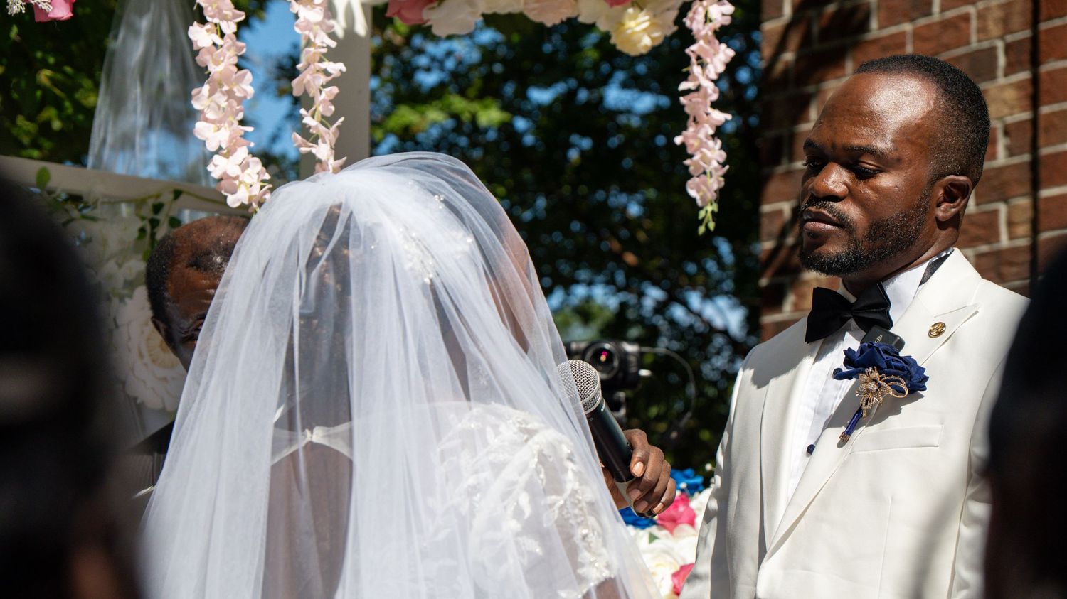 A groom in a white suit holds his veiled bride's hand during an outdoor wedding ceremony.