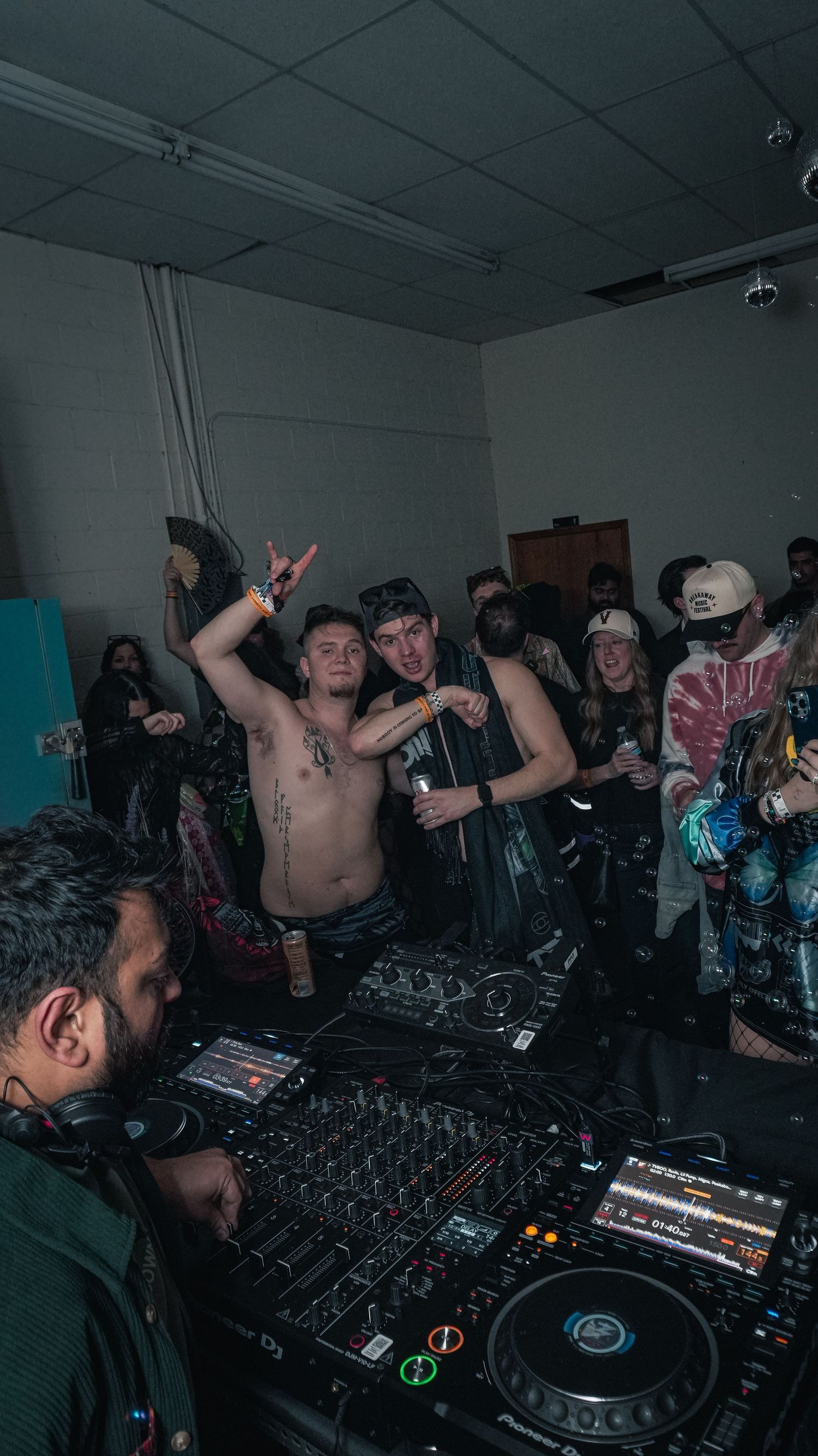 Shirtless partygoers posing near DJ equipment at an underground music event.