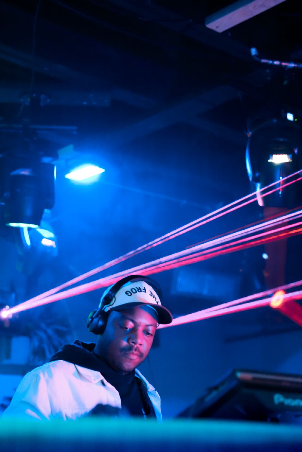 DJ wearing headphones and cap performing under red laser lights in a dark nightclub.