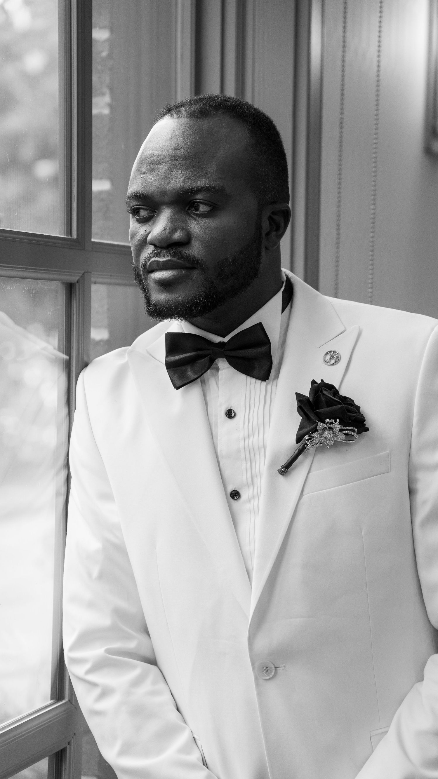 An elegant groom in a white tuxedo with a black bow tie poses in black and white.
