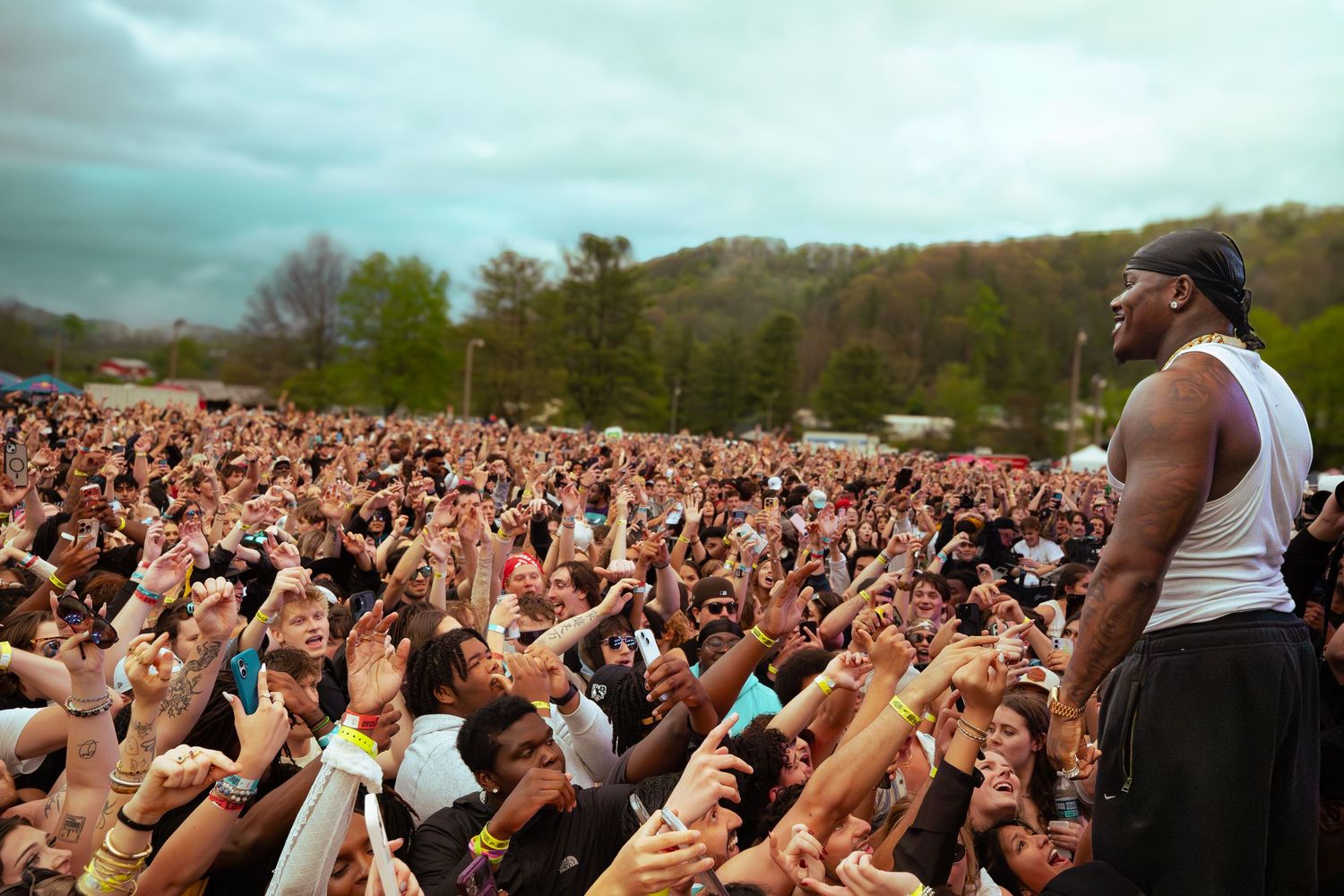 A performer in a white tank top stands before a huge crowd at an outdoor music festival.