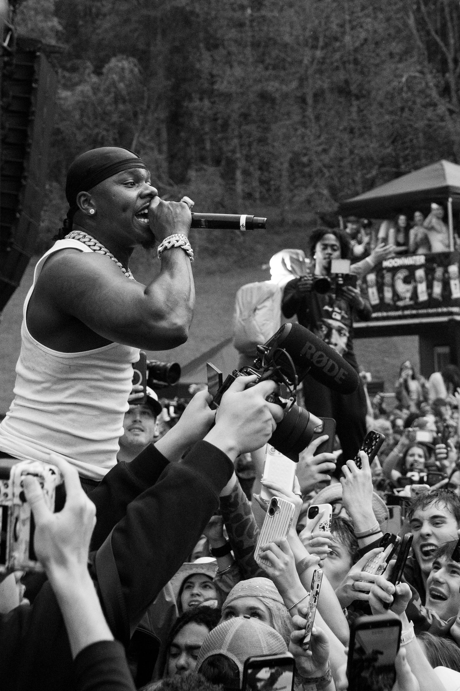 Black and white photo of male rapper performing into mic above enthusiastic crowd at outdoor festival.