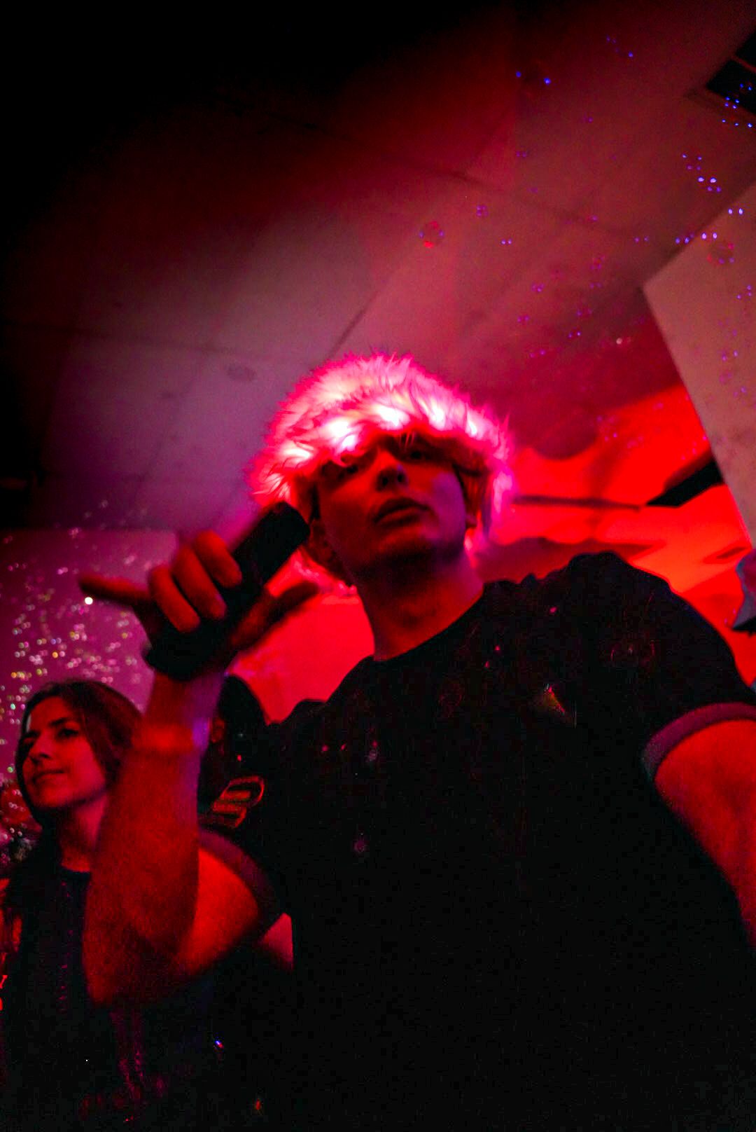 Performer holding microphone wearing a fluffy pink hat under dramatic red club lighting.