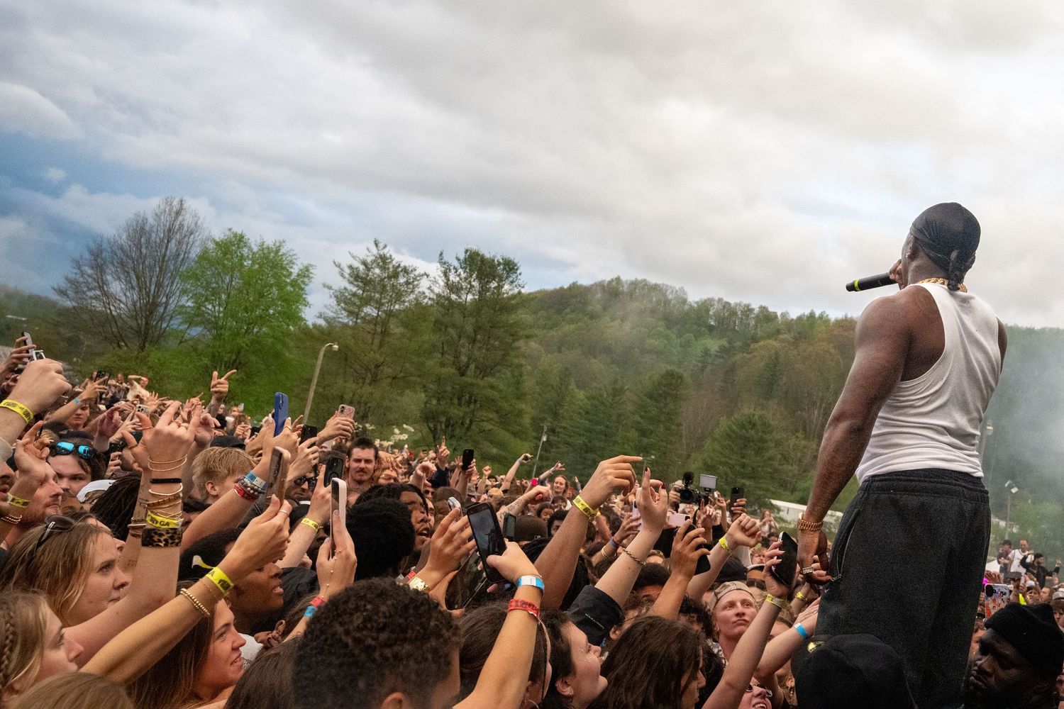 Male rapper in white tank top performs atop crowd at outdoor music festival with green hills behind.