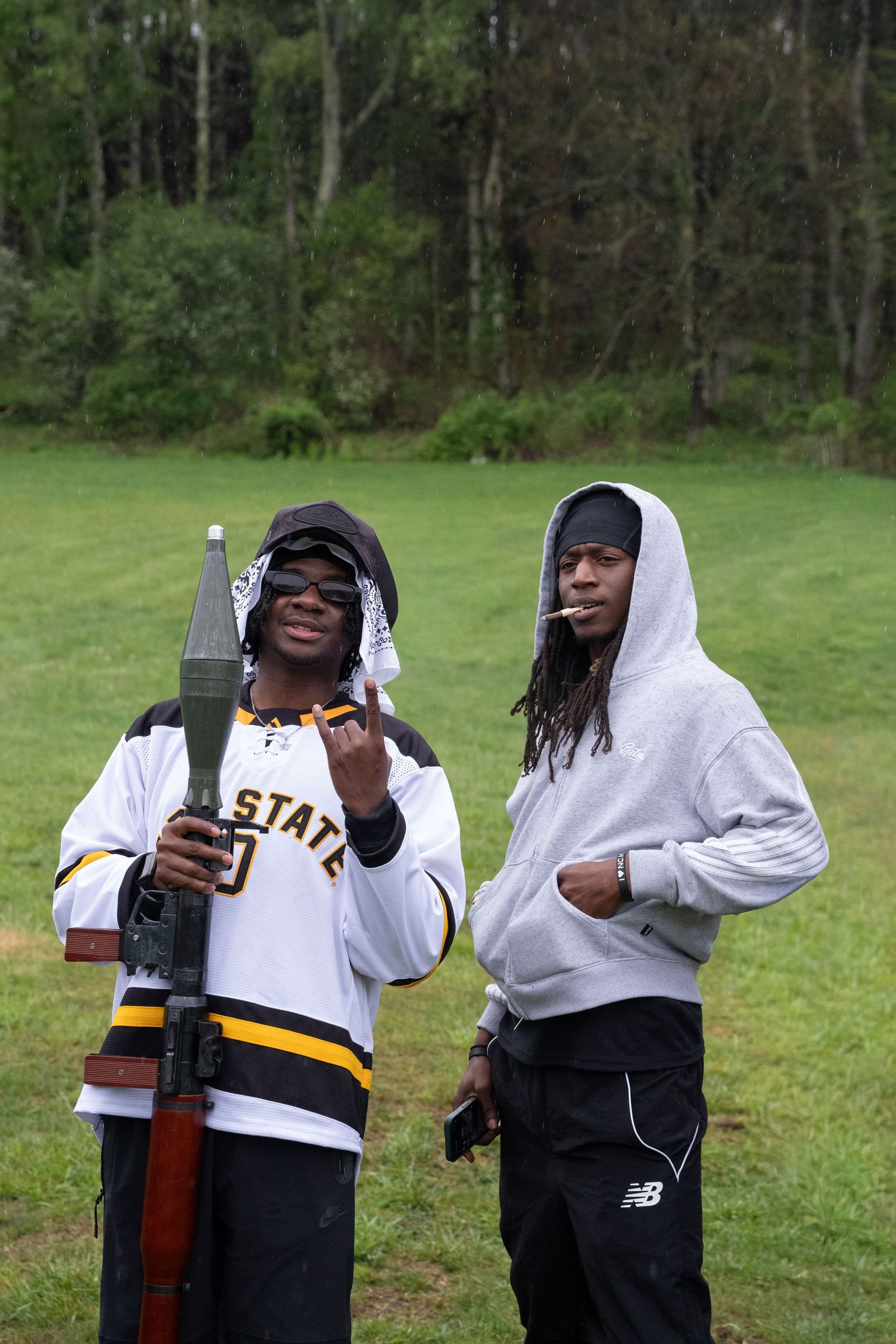 Two men pose outdoors, one holding a prop rifle, at a casual outdoor gathering.