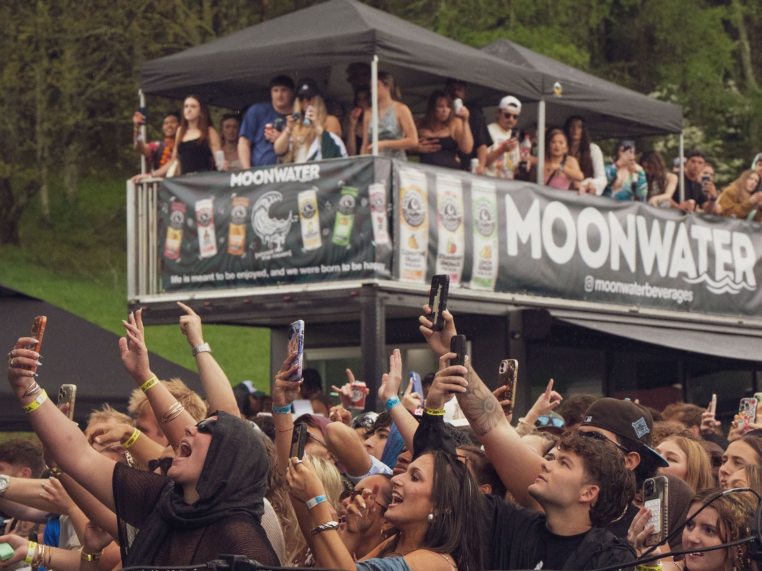 Large crowd filming with phones near Moonwater branded booth at outdoor music festival.